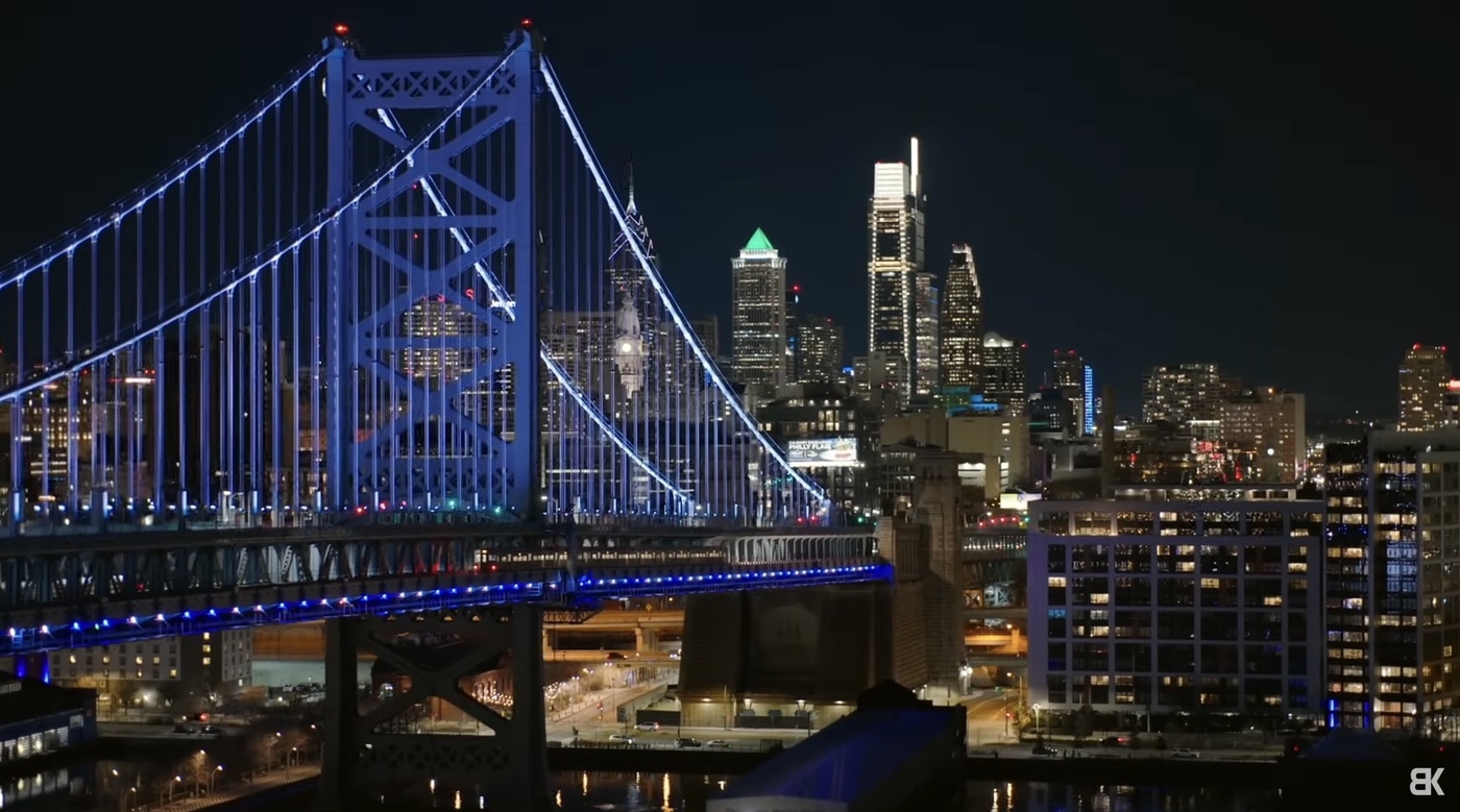 Night aerial of the blue-lit Benjamin Franklin Bridge and the Philadelphia skyline.