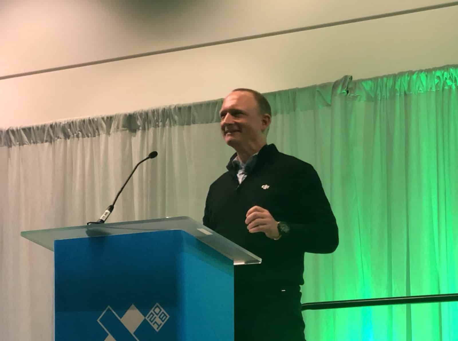 A speaker in a black DJI polo addresses a conference from a green-lit lectern.