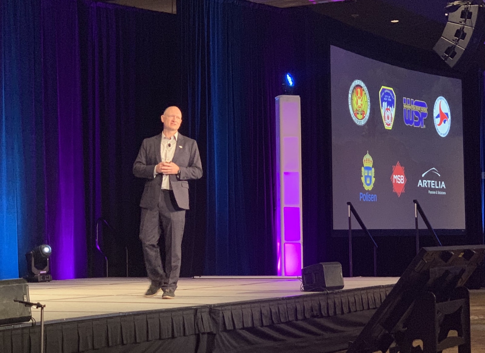 A keynote speaker stands beside a screen filled with public safety agency badges.