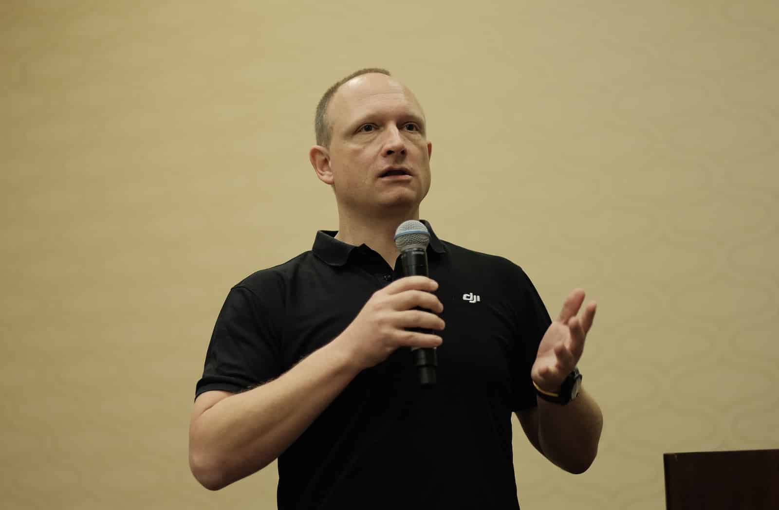 A DJI staffer holds a microphone mid-sentence while presenting to a seated audience.