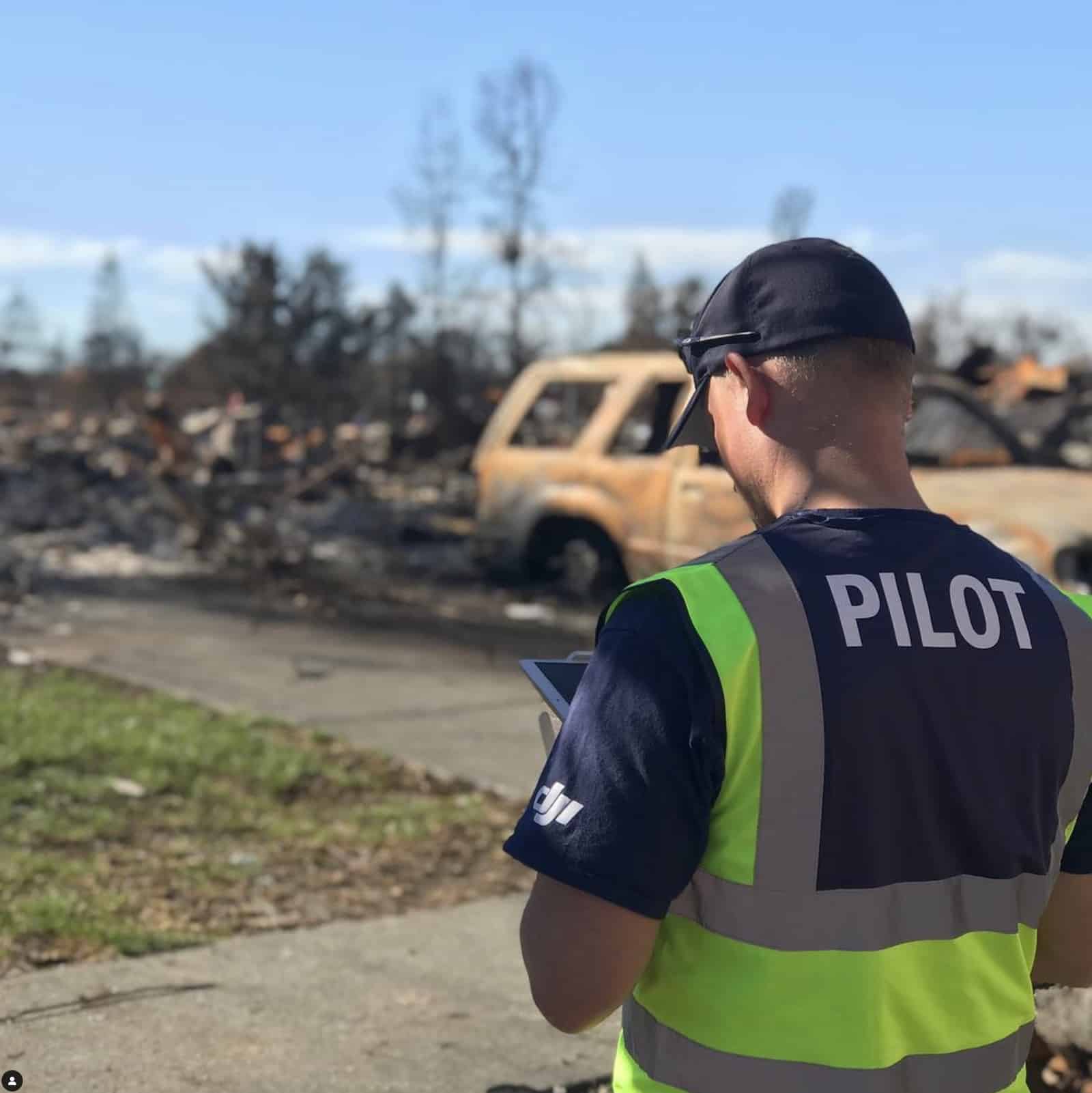 A pilot in a high-visibility vest reviews a tablet amid burned-out wildfire wreckage.