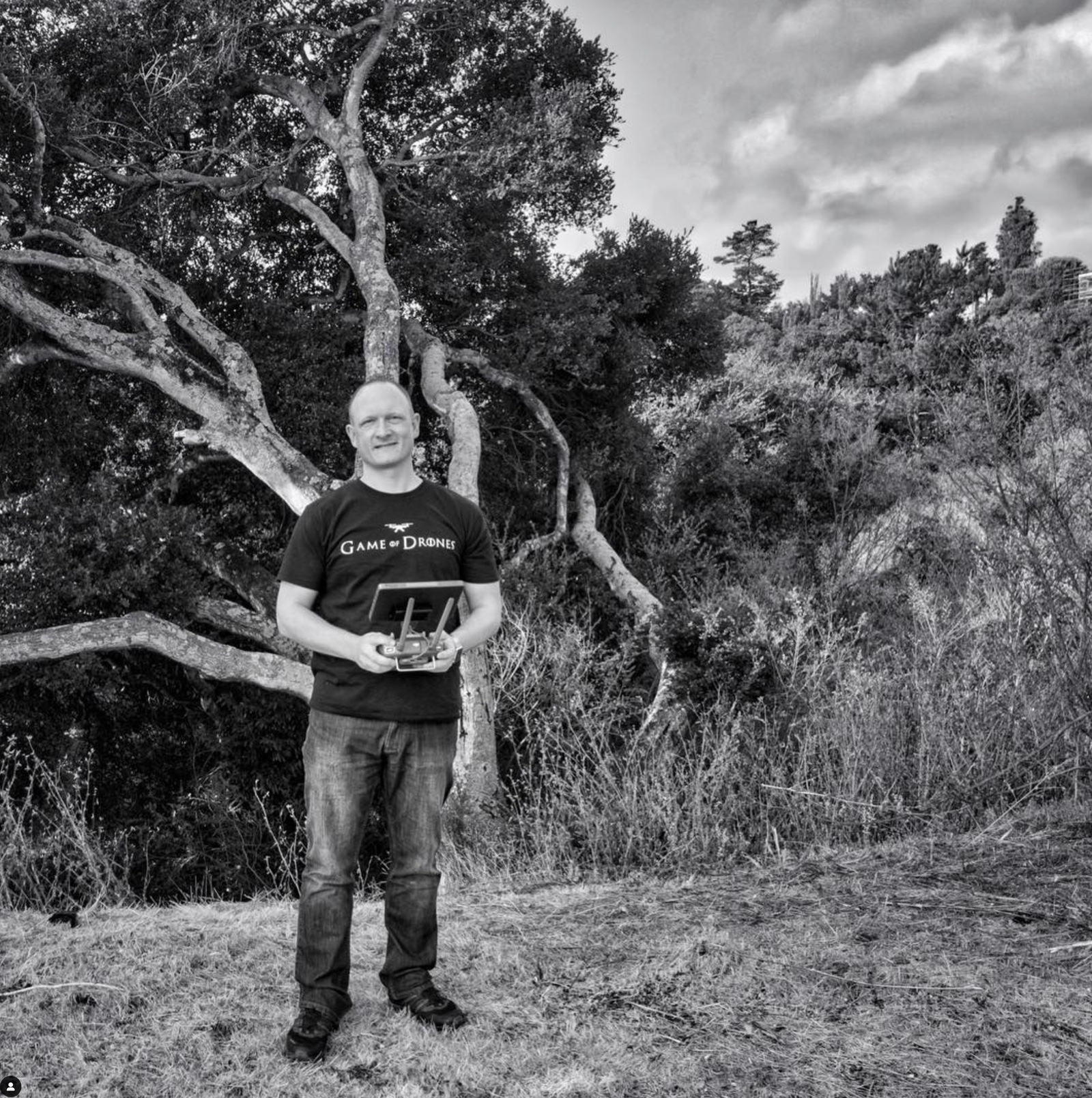 Monochrome portrait of a pilot holding a controller beneath a gnarled hillside tree.
