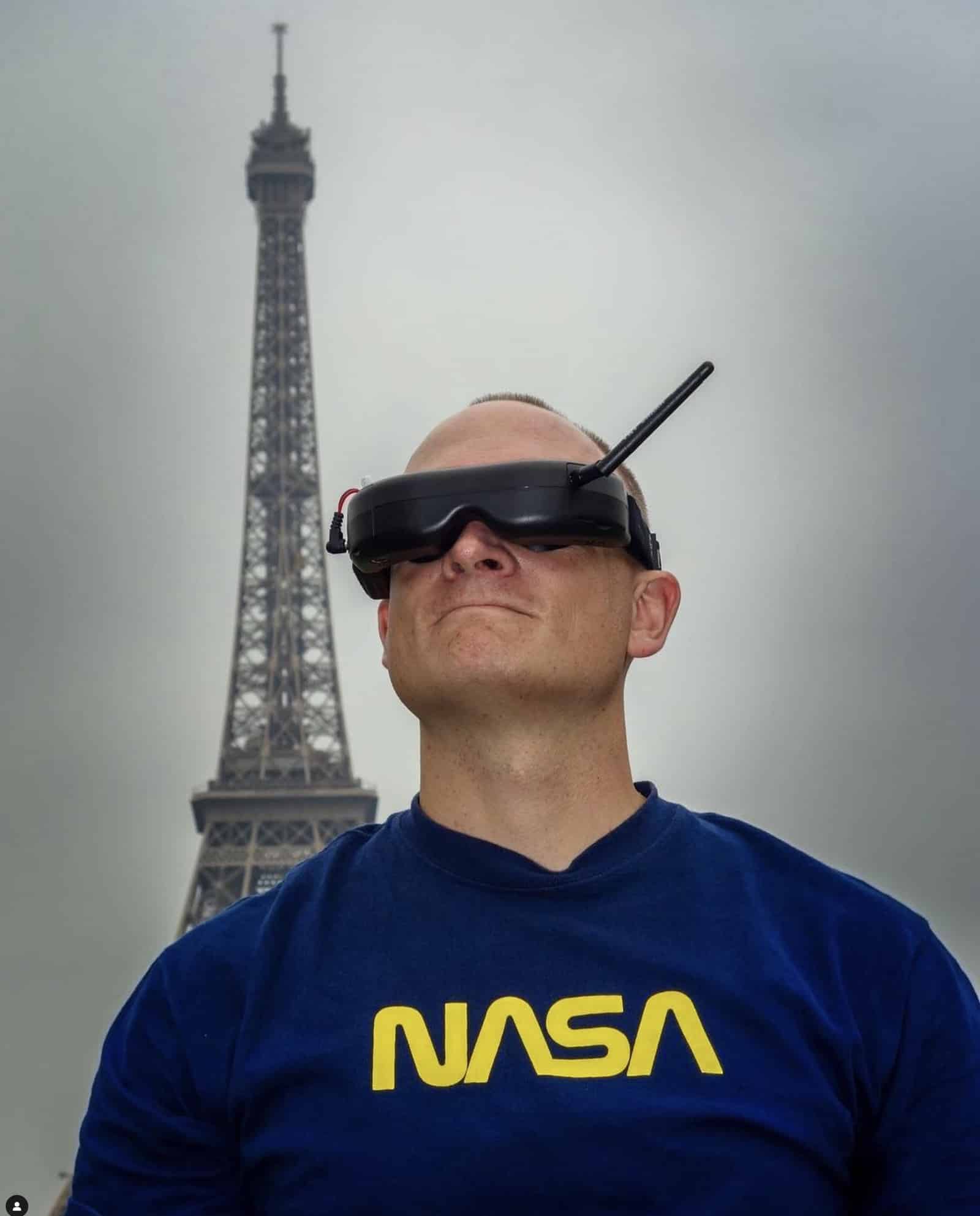 A man in a NASA sweatshirt wears FPV goggles before an Eiffel Tower backdrop.