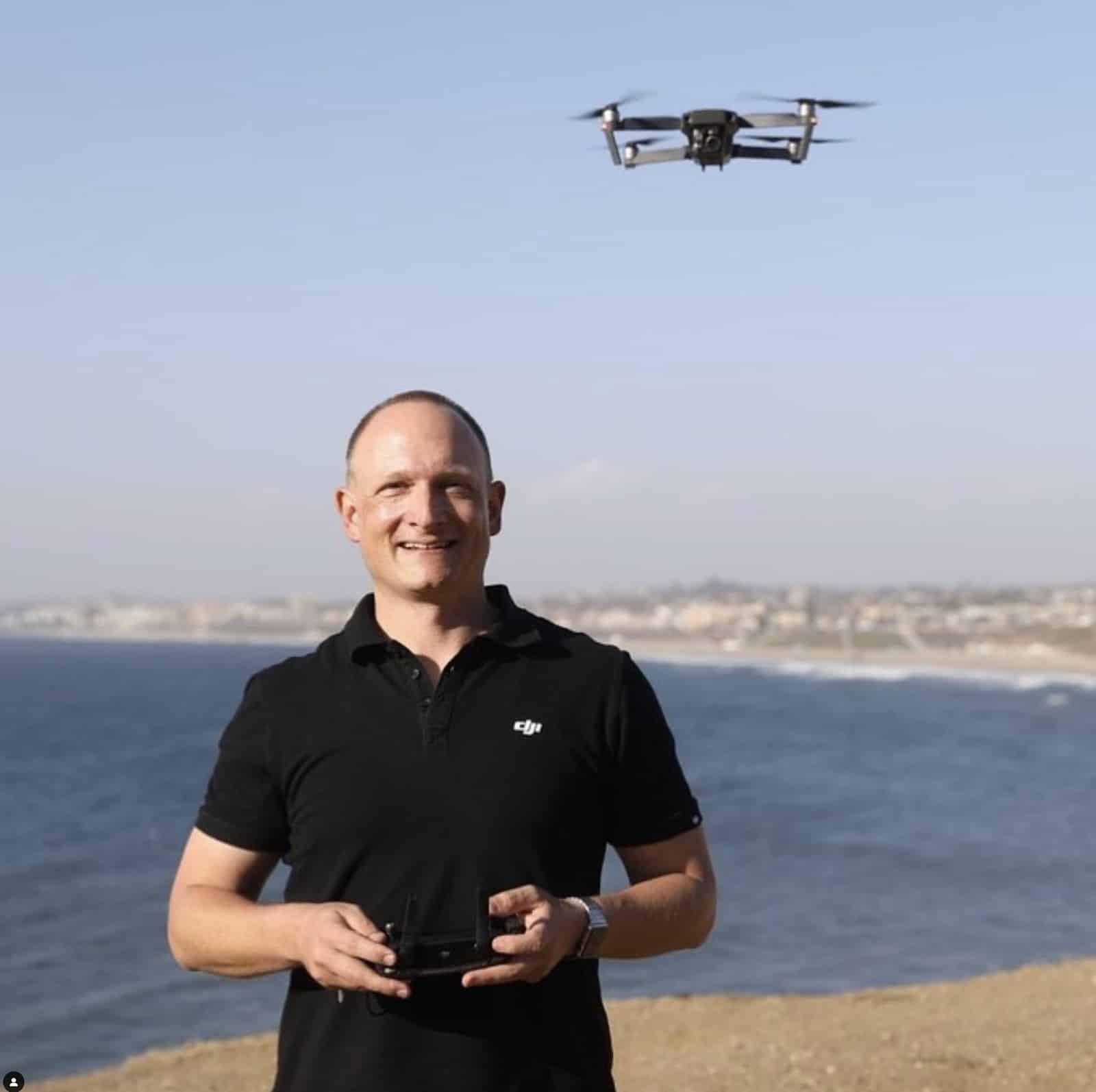 A pilot on a coastal bluff flies a folding drone hovering overhead.