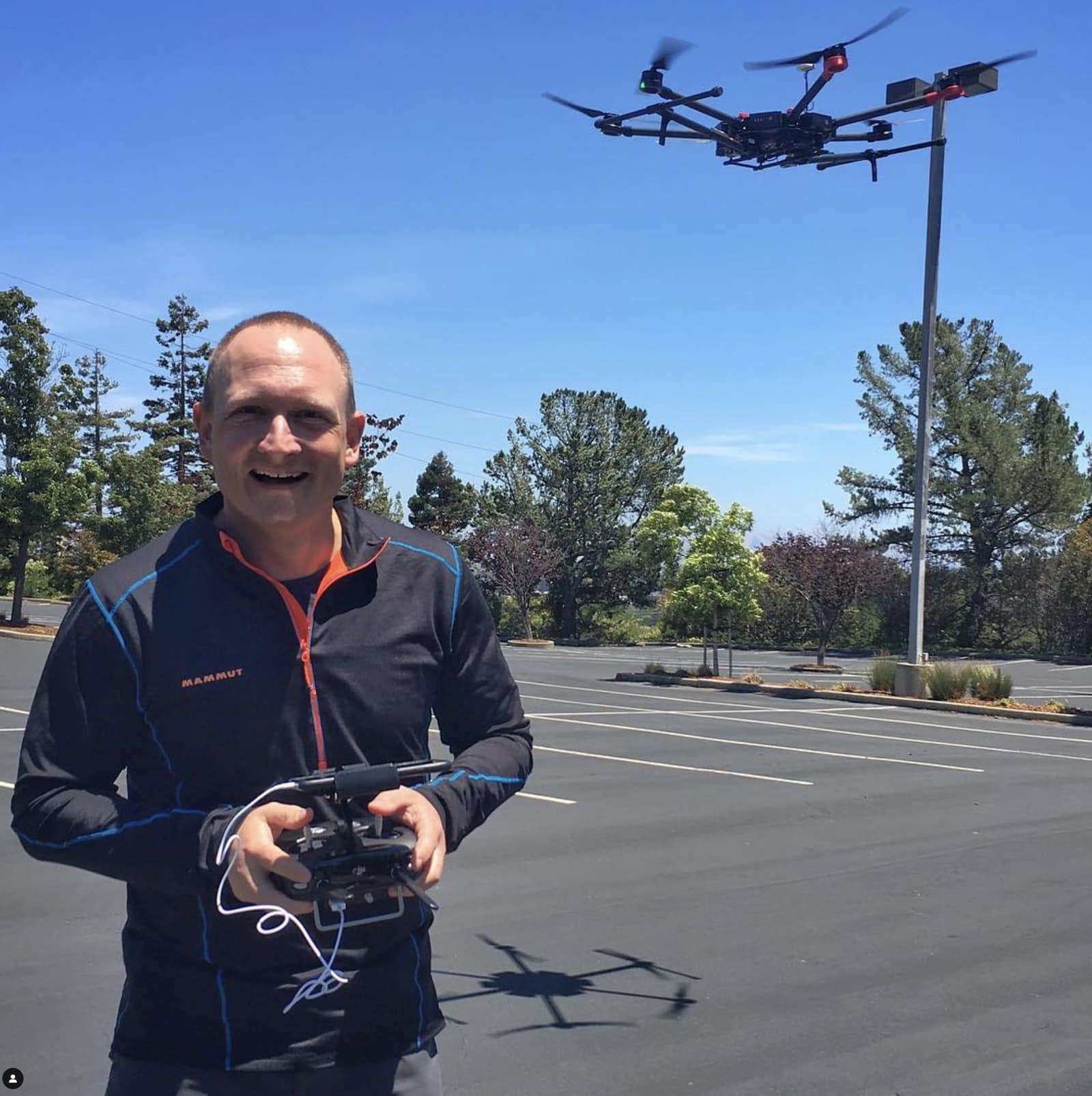 A pilot in a blue jacket flies a hexacopter above an empty parking lot.
