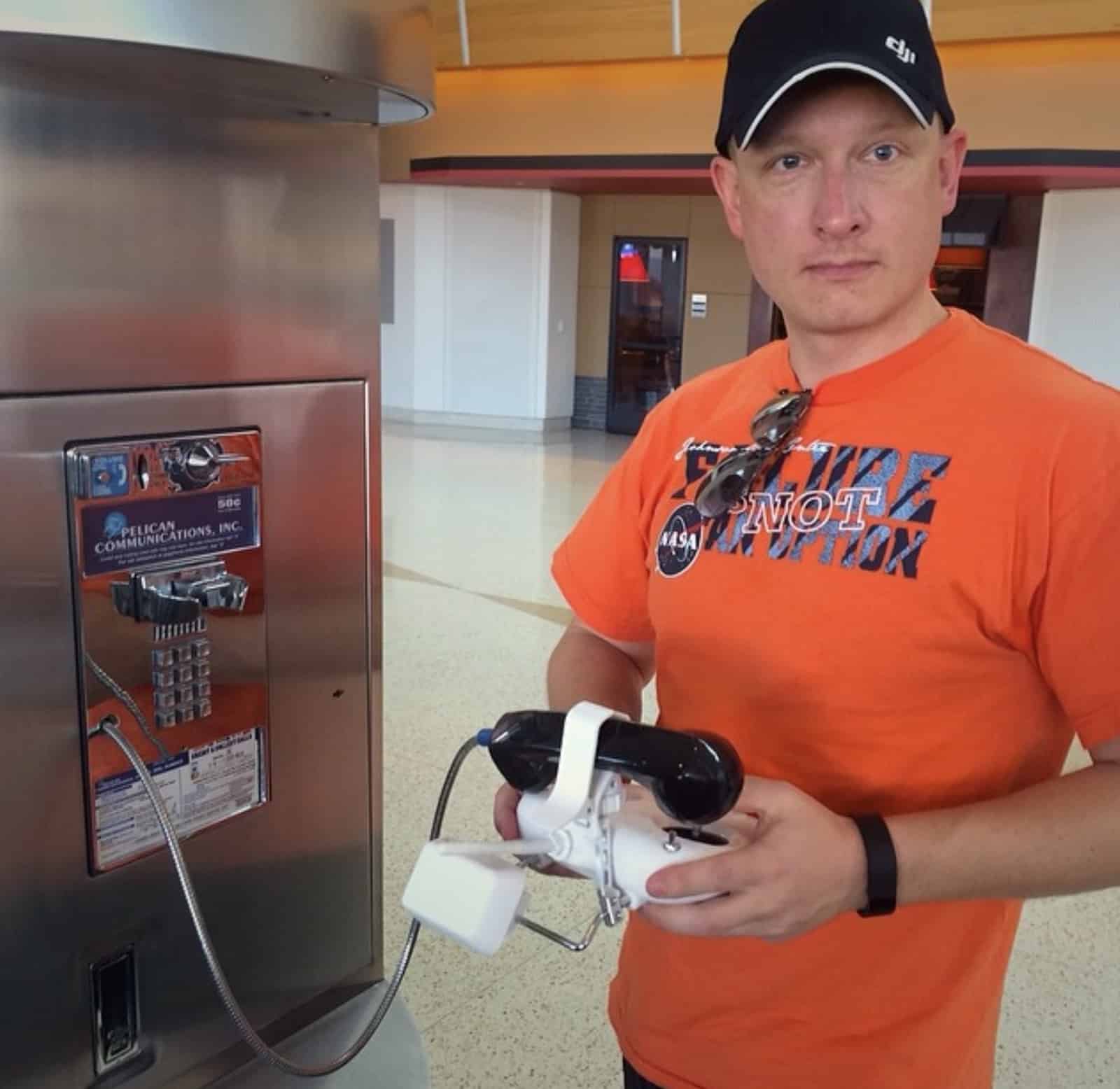 A man in a DJI cap holds a white drone controller beside a payphone.