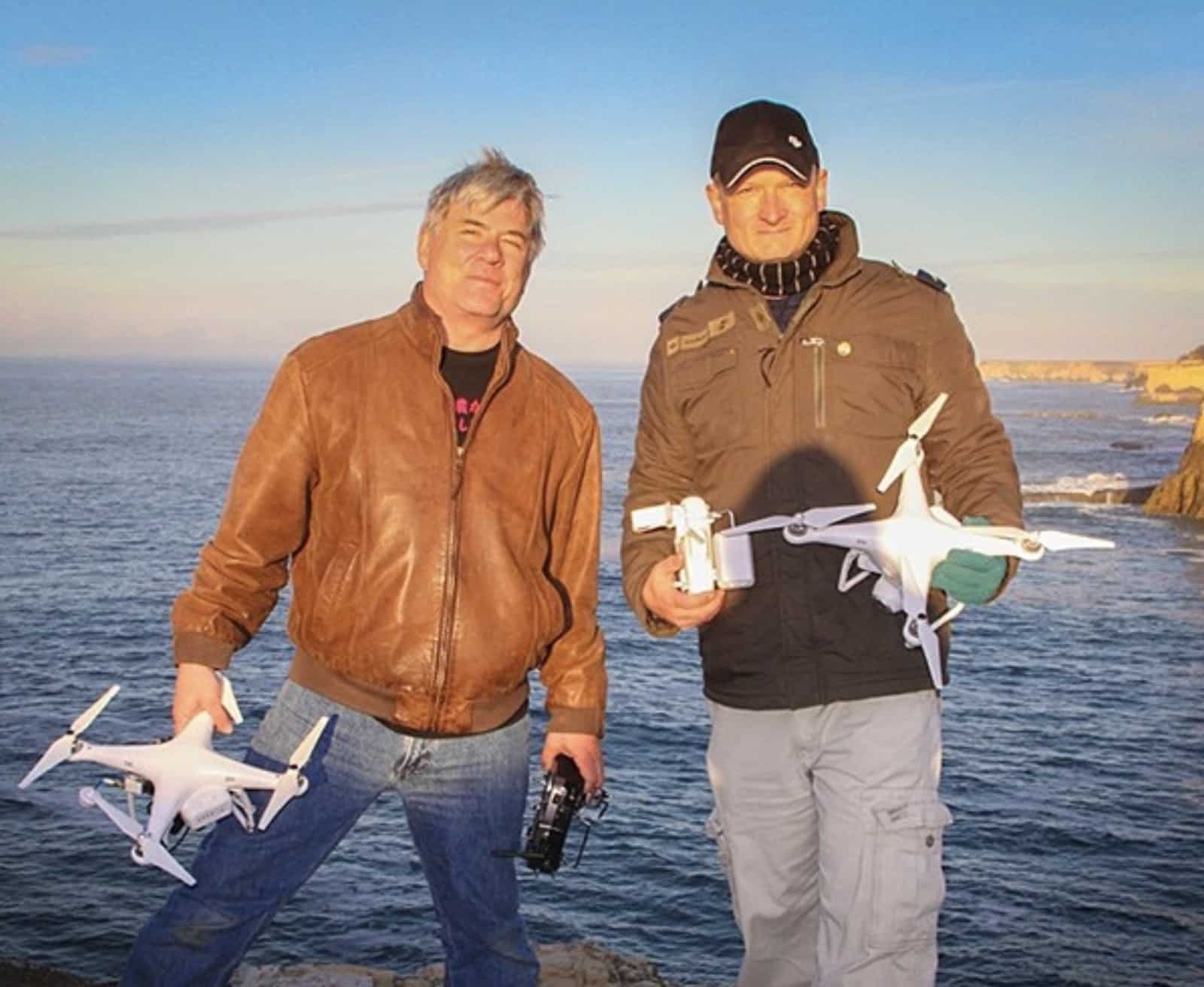 Two pilots hold DJI Phantom quadcopters on a cliff above the ocean.