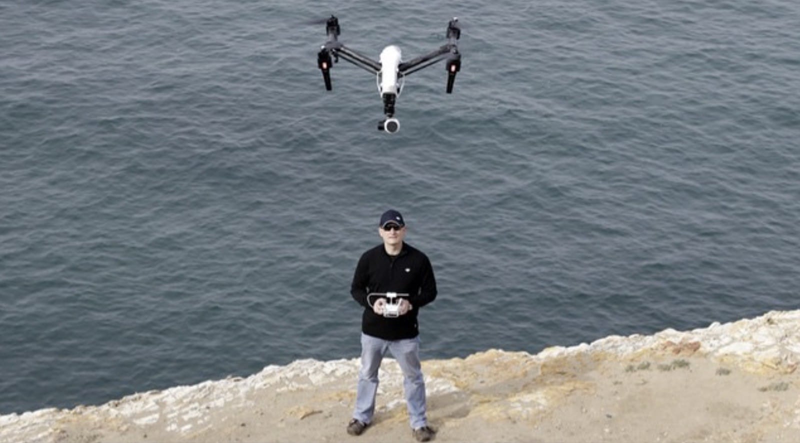 A pilot on a rocky shoreline flies a DJI Inspire hovering directly above him.