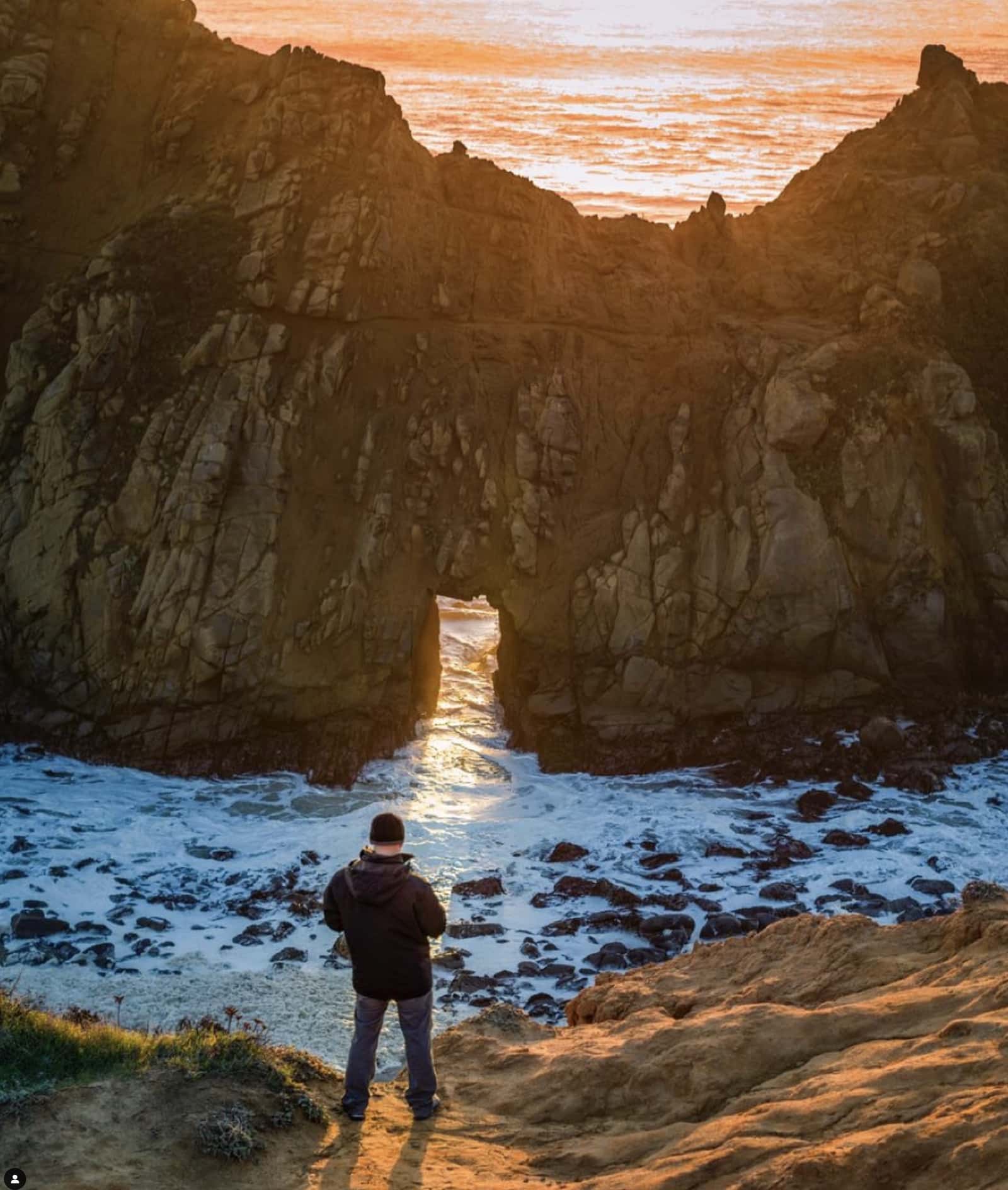 A lone figure watches sunset light stream through a coastal sea arch.