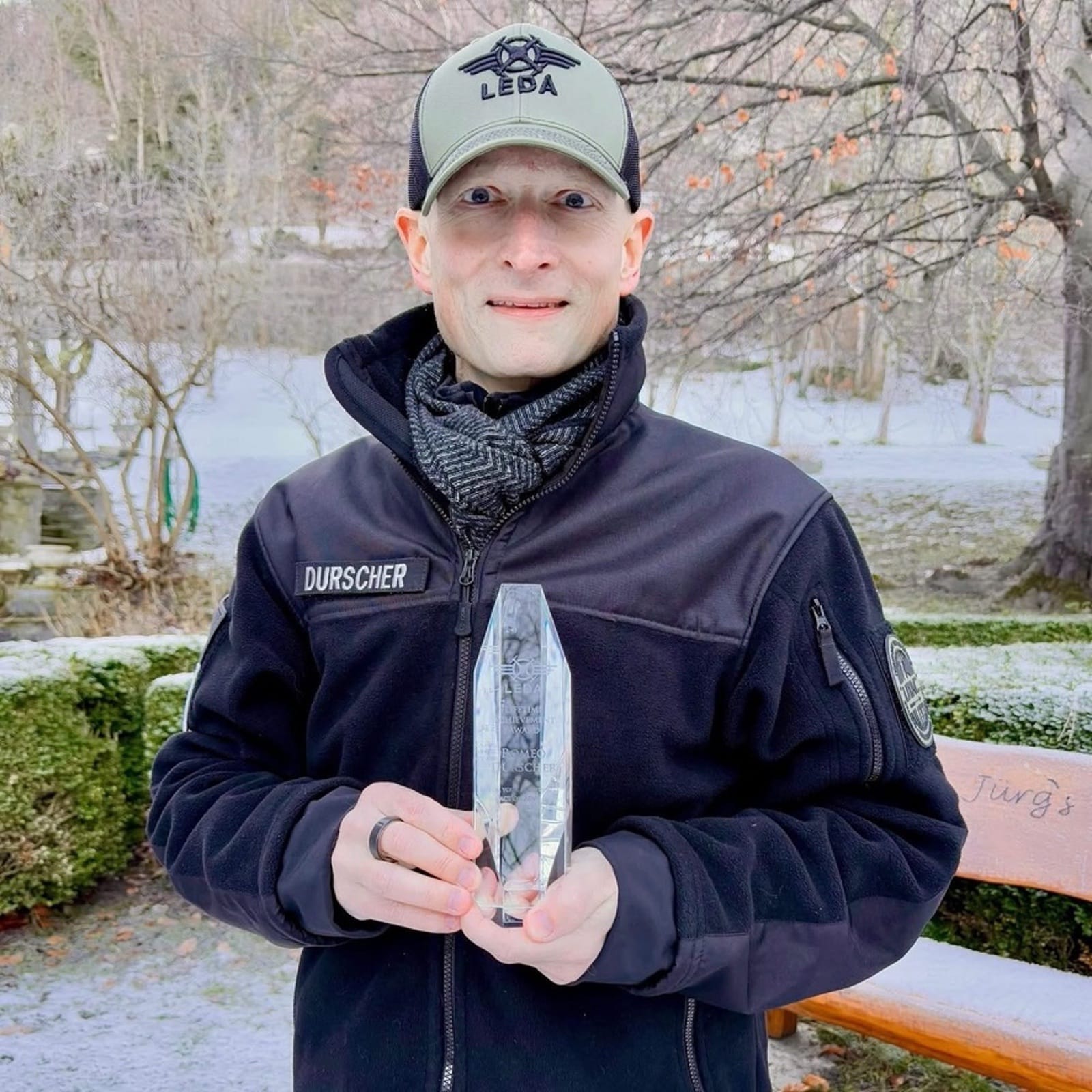 A man in a jacket with a Durscher nametape holds a glass trophy outdoors.
