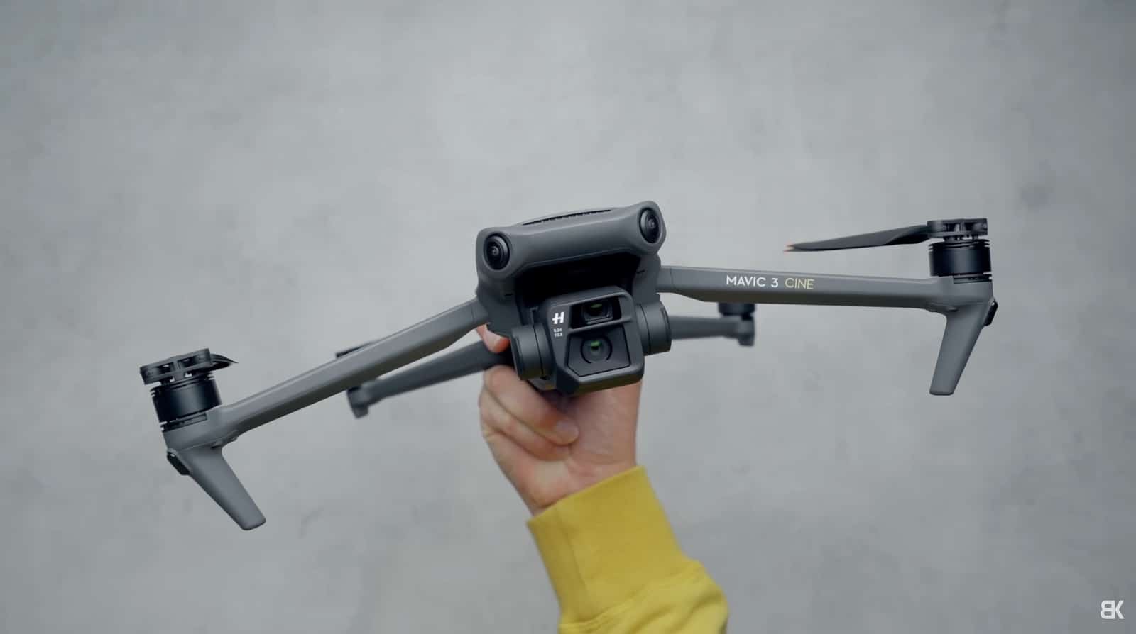 A hand in a yellow sleeve holds a Mavic 3 Cine against a gray wall.