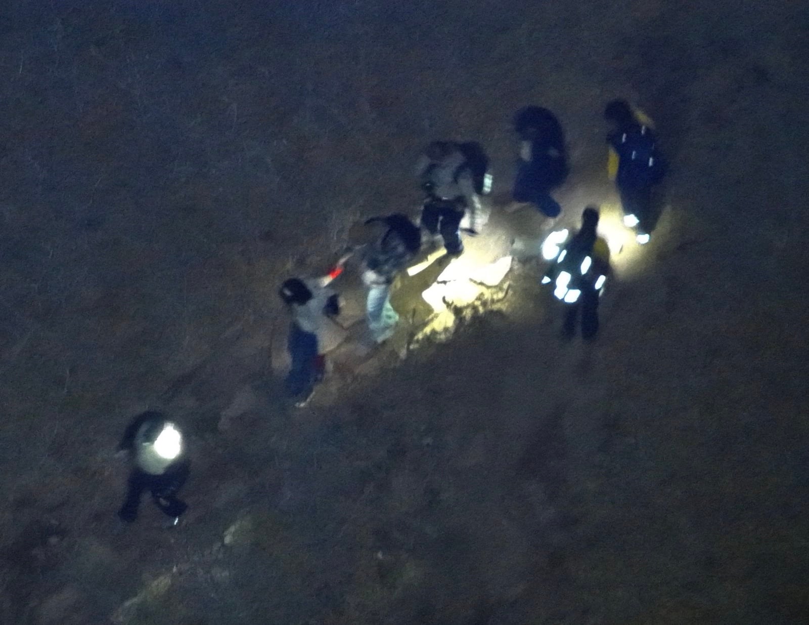 Aerial night view of a rescue team with headlamps gathered on a hillside.
