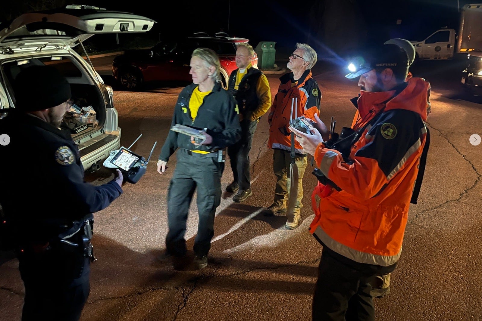 Rescue crews hold drone controllers at a trailhead staging area after dark.