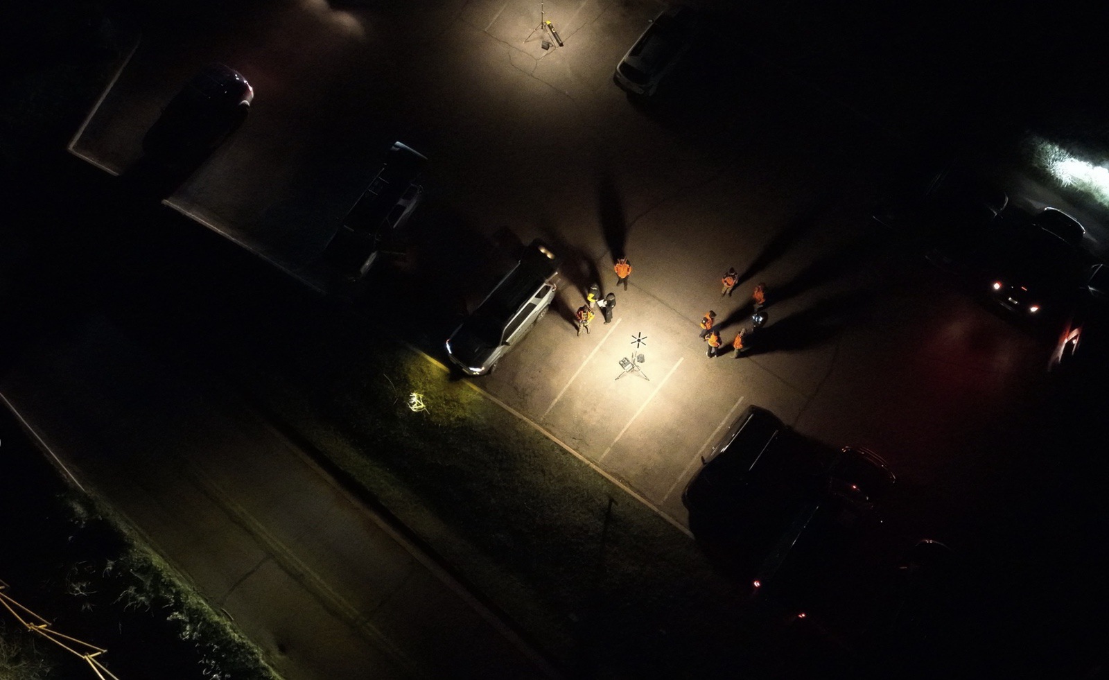 Overhead night view of vehicles and searchers lit by a drone spotlight.