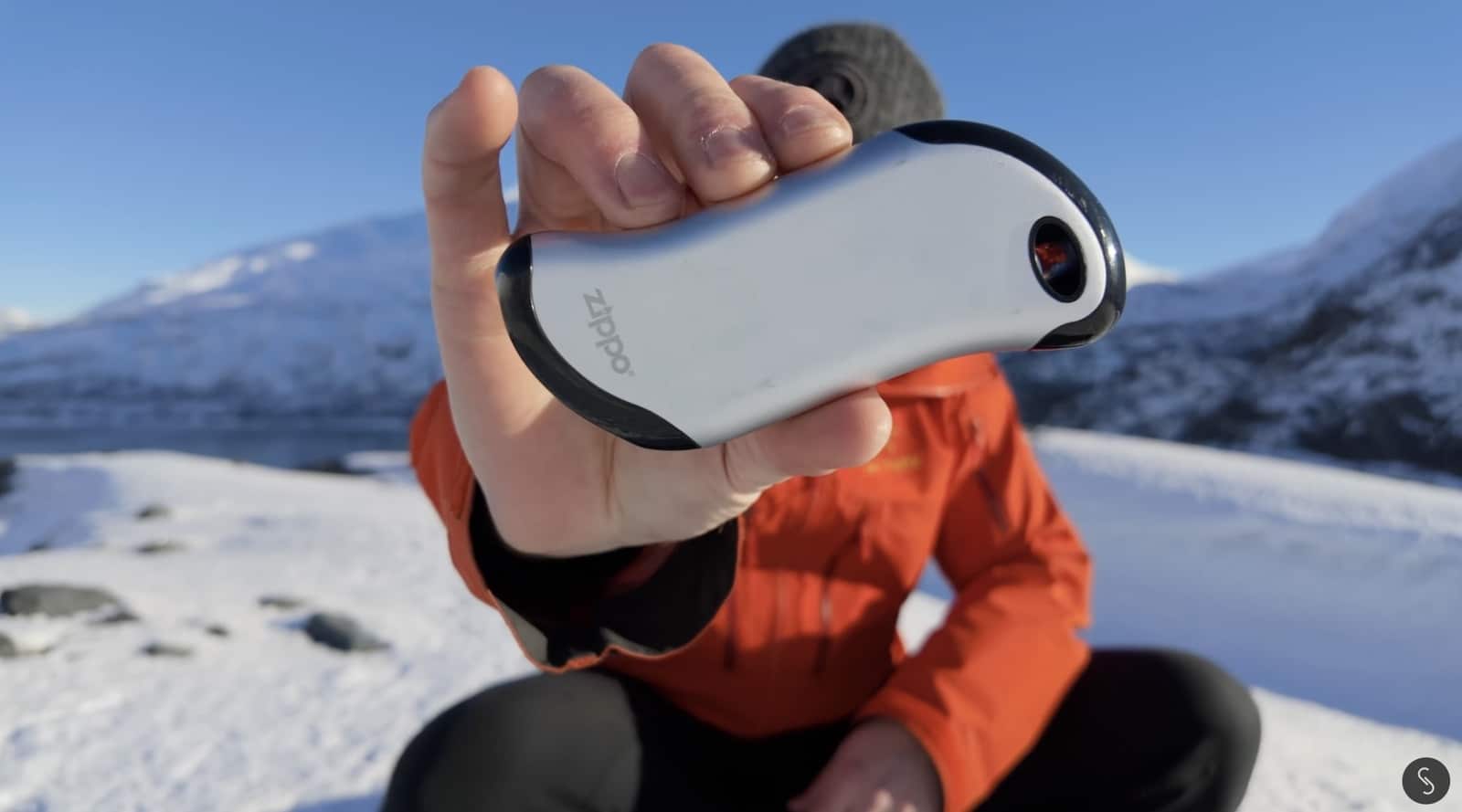 Bare hand holding a white Zippo hand warmer above a snow-covered slope.