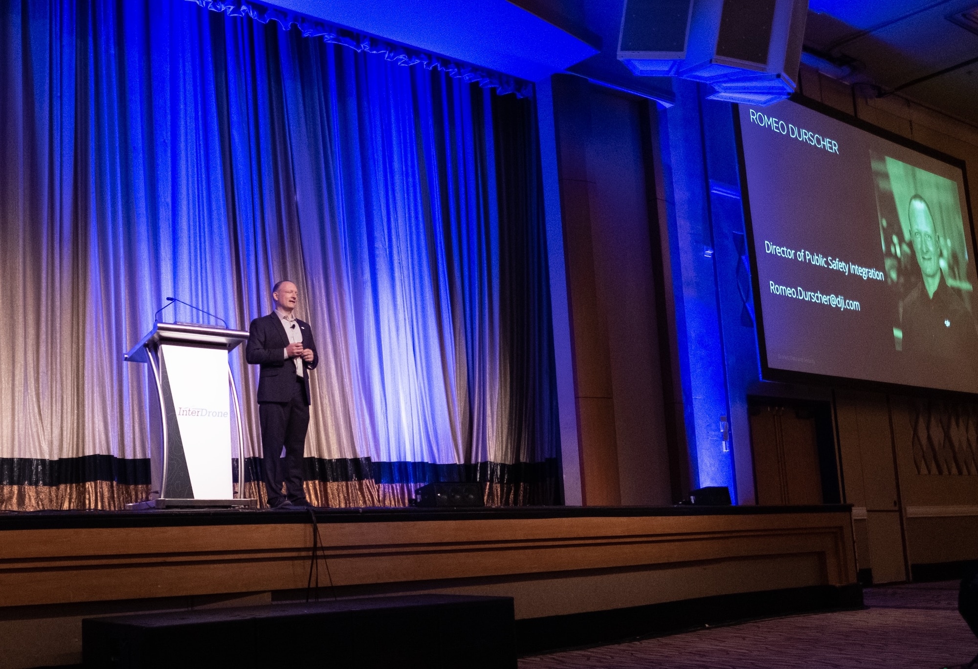 Romeo Durscher speaks from a lectern at InterDrone in 2018 under blue stage lighting.