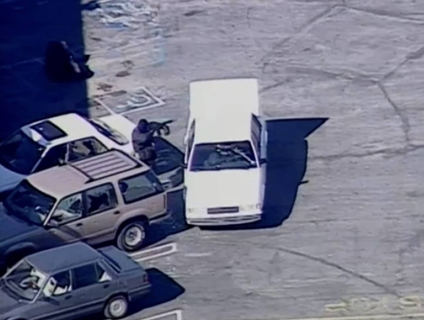 Grainy aerial news frame of an armed standoff among parked cars.