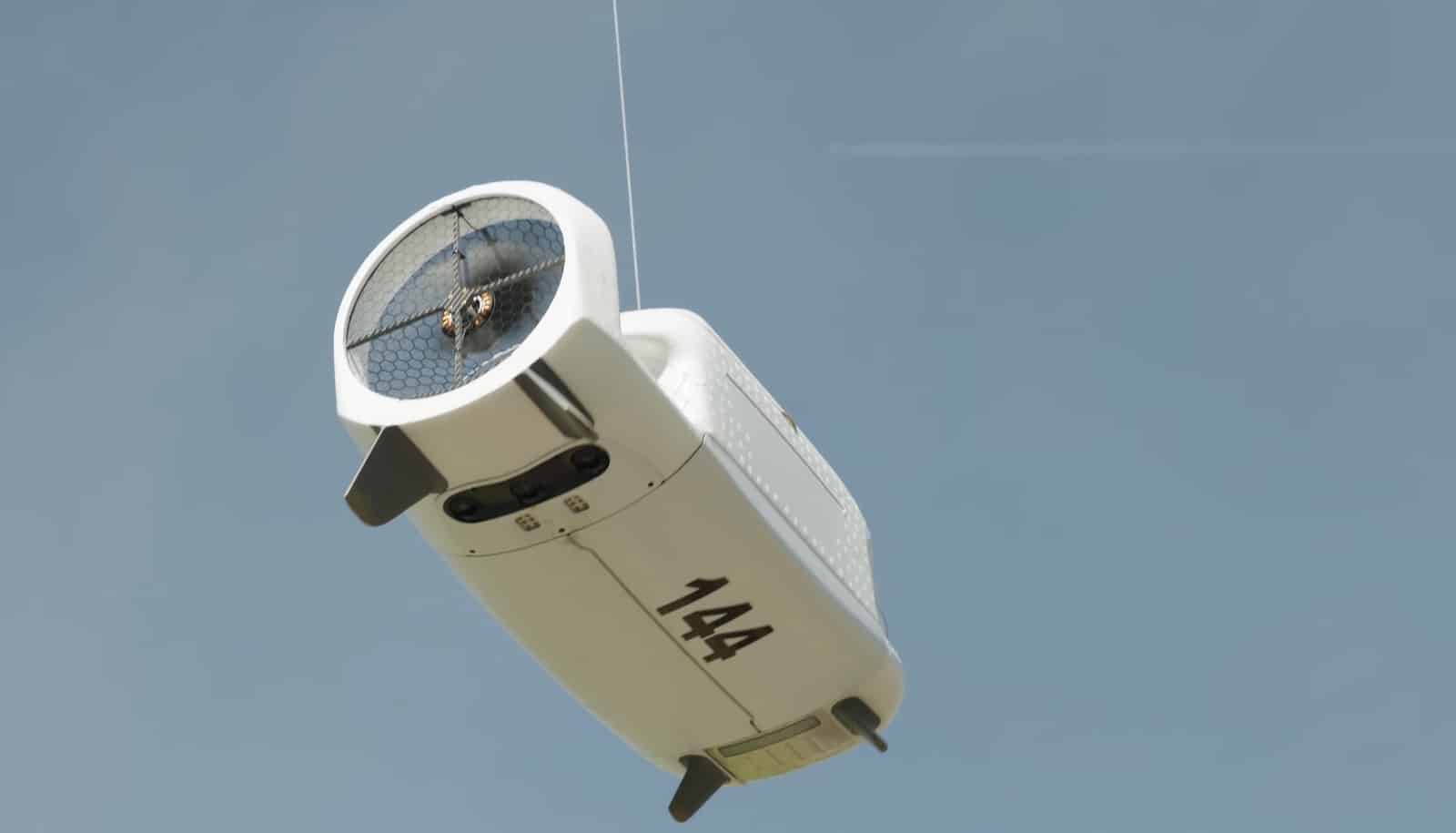 Zipline delivery droid numbered 144 descends on its tether against a clear blue sky.