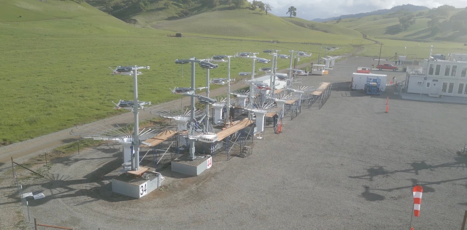 A row of docking masts lines a gravel pad below green rolling hills.