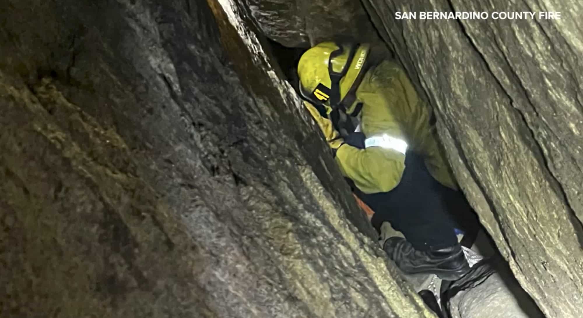 A firefighter in yellow turnout gear works head-down inside a narrow granite crevice.