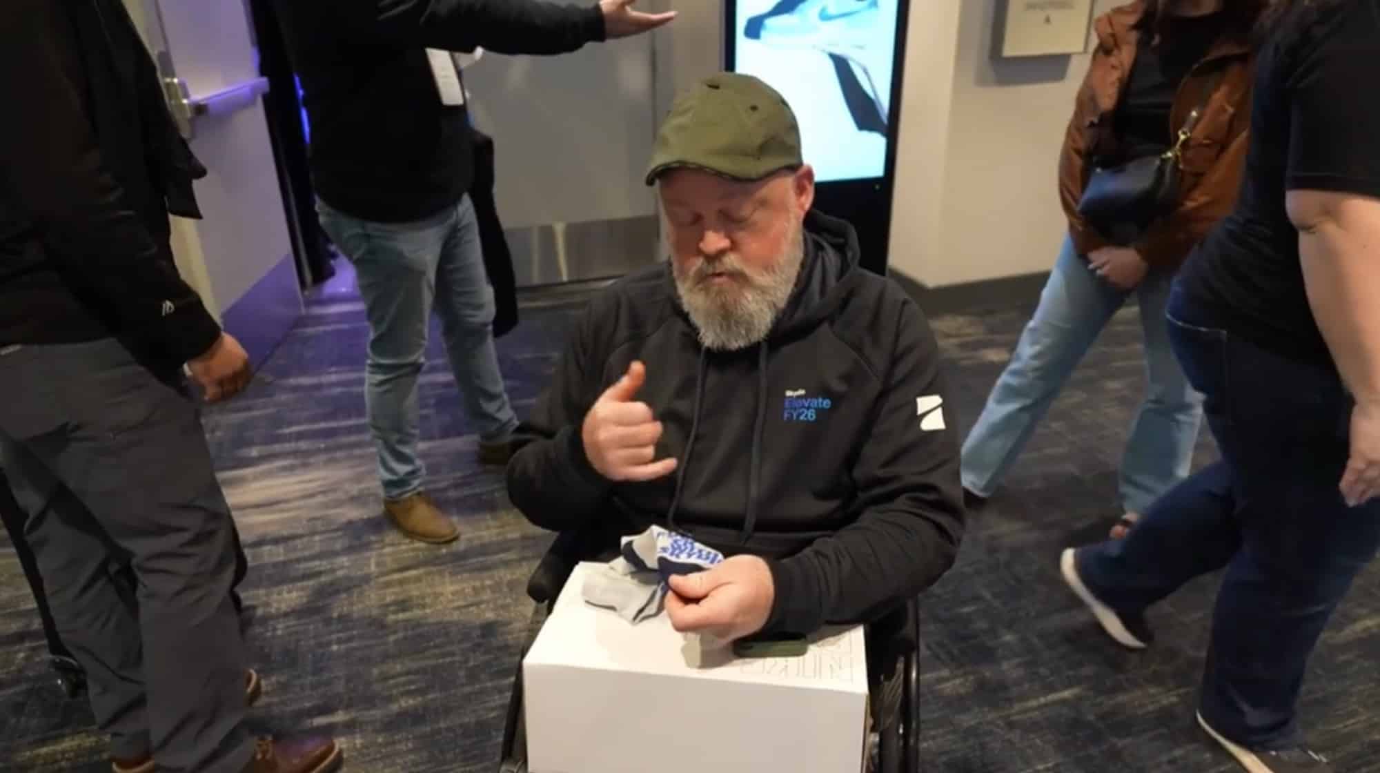 Bearded attendee in a cap gives a thumbs up from a white conference stool.