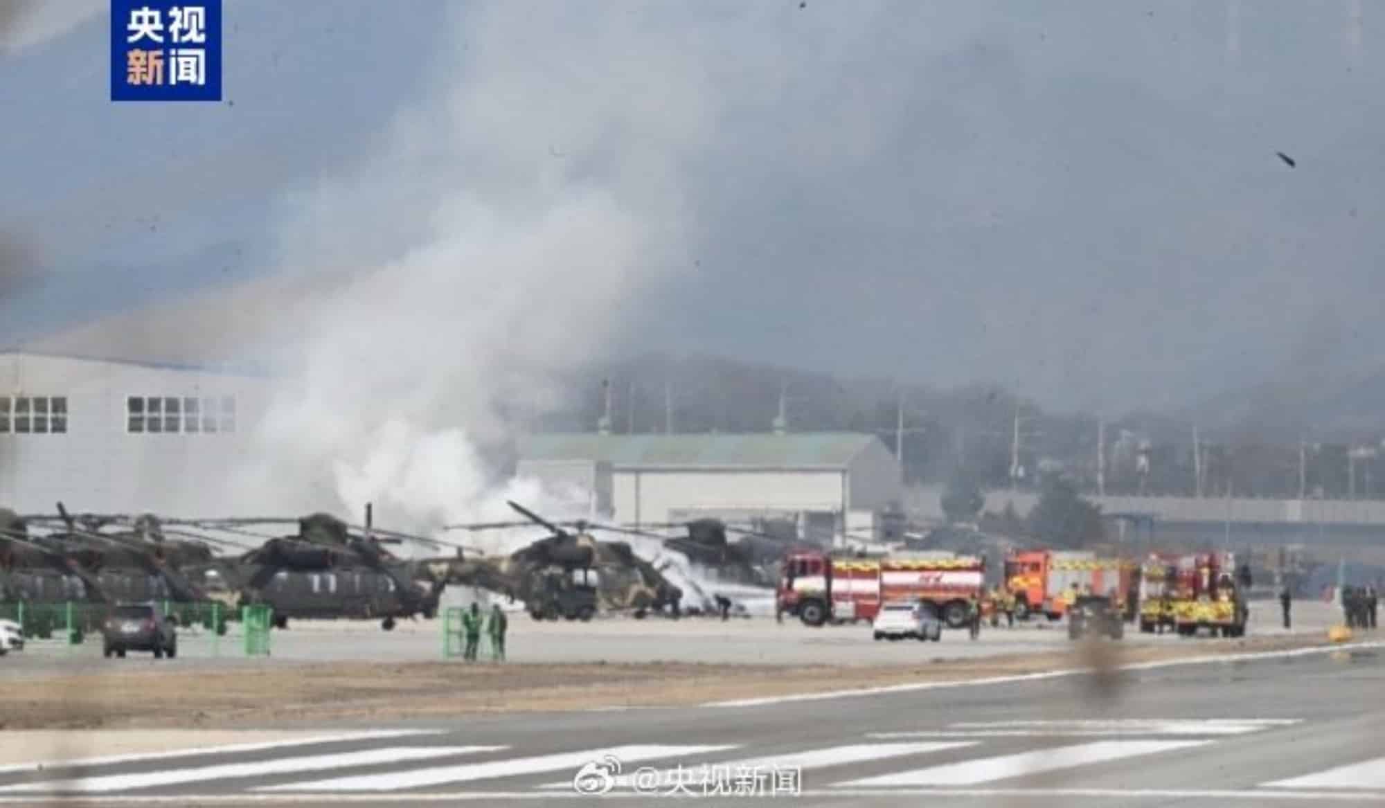 Broadcast still of smoke rising over an airfield lined with helicopters and fire trucks.