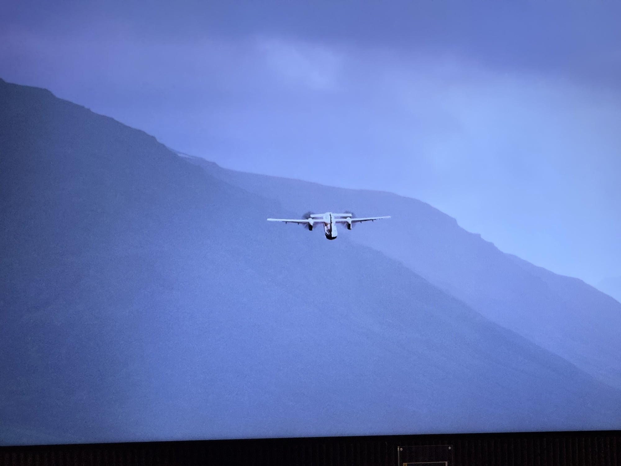 A transport aircraft flies past a dark hillside in projected conference footage.