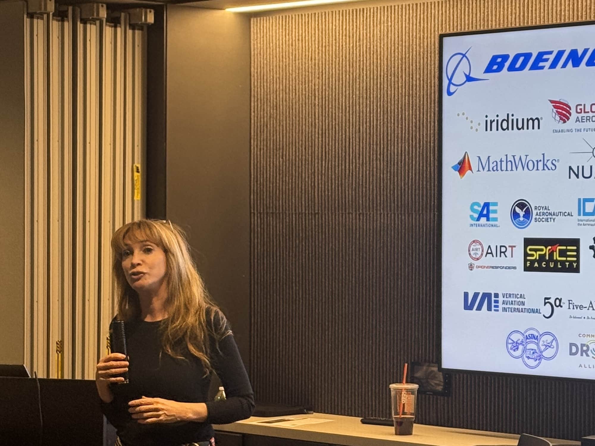 A speaker addresses the room beside a screen listing GoAERO sponsor logos.