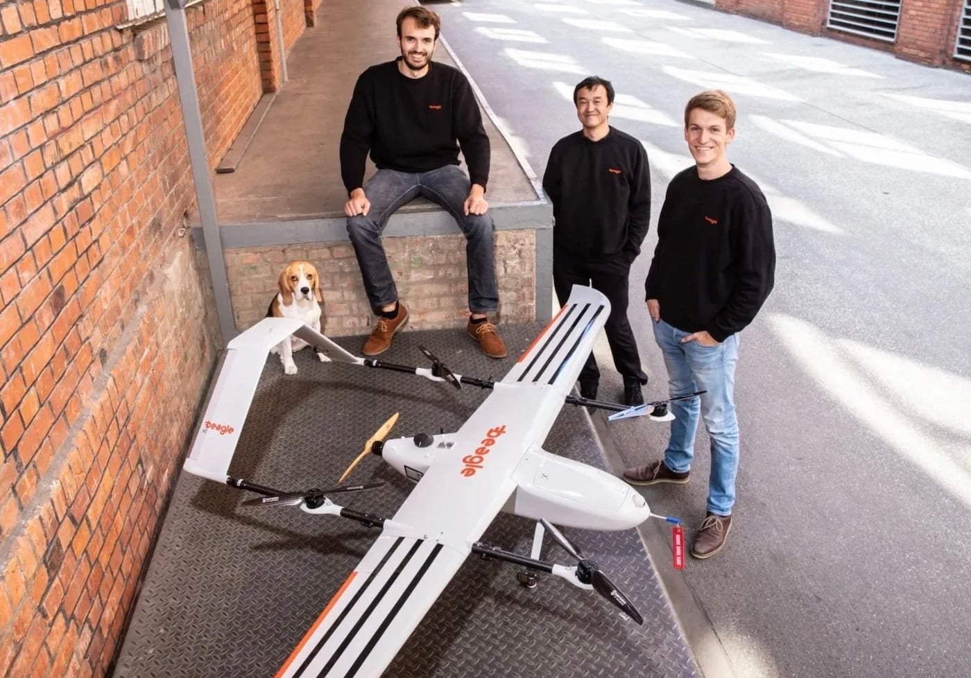 Three Beagle co-founders and a beagle dog pose beside their fixed-wing VTOL drone.