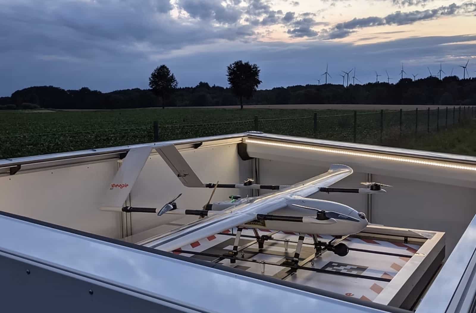 Long-range Beagle drone sits inside an open docking station at dusk near wind turbines.