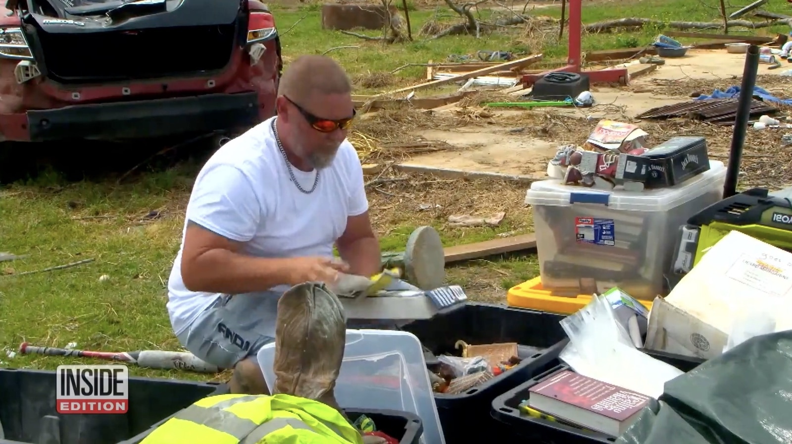 Man kneeling among salvaged bins and storm debris in a wrecked yard.