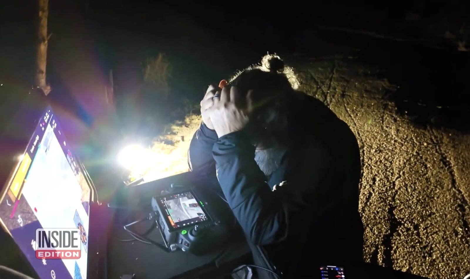Night frame of a drone pilot at a lit controller beside a dirt bank.