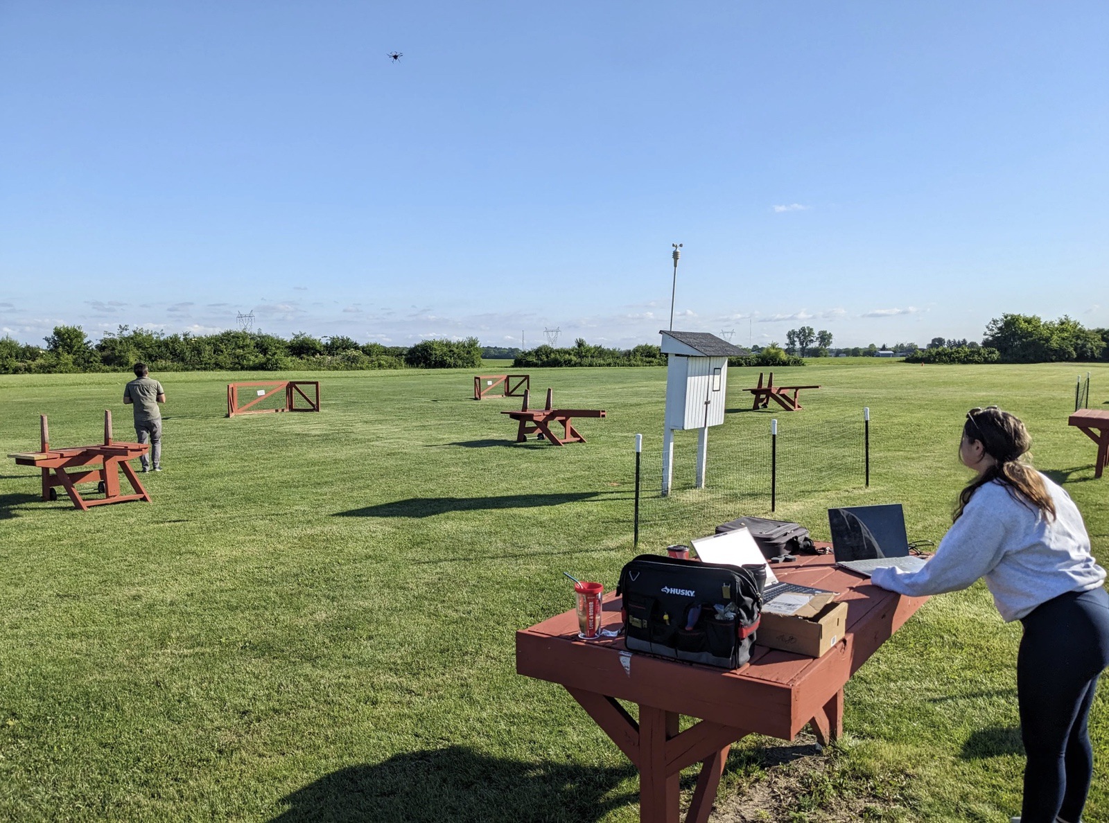 Researcher works at a laptop on a field test range as a drone flies overhead.