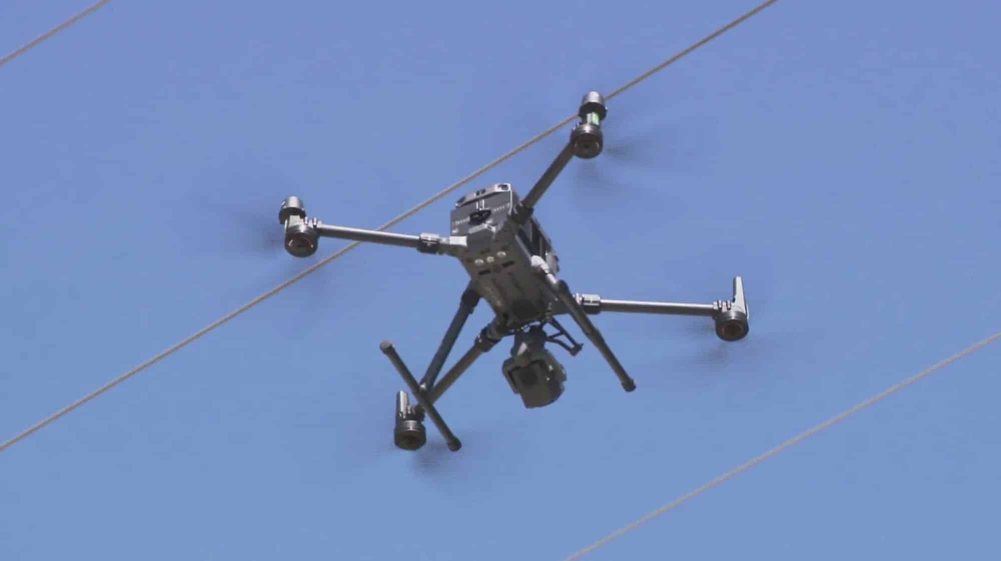 A drone hovers beside an overhead power line against clear blue sky.