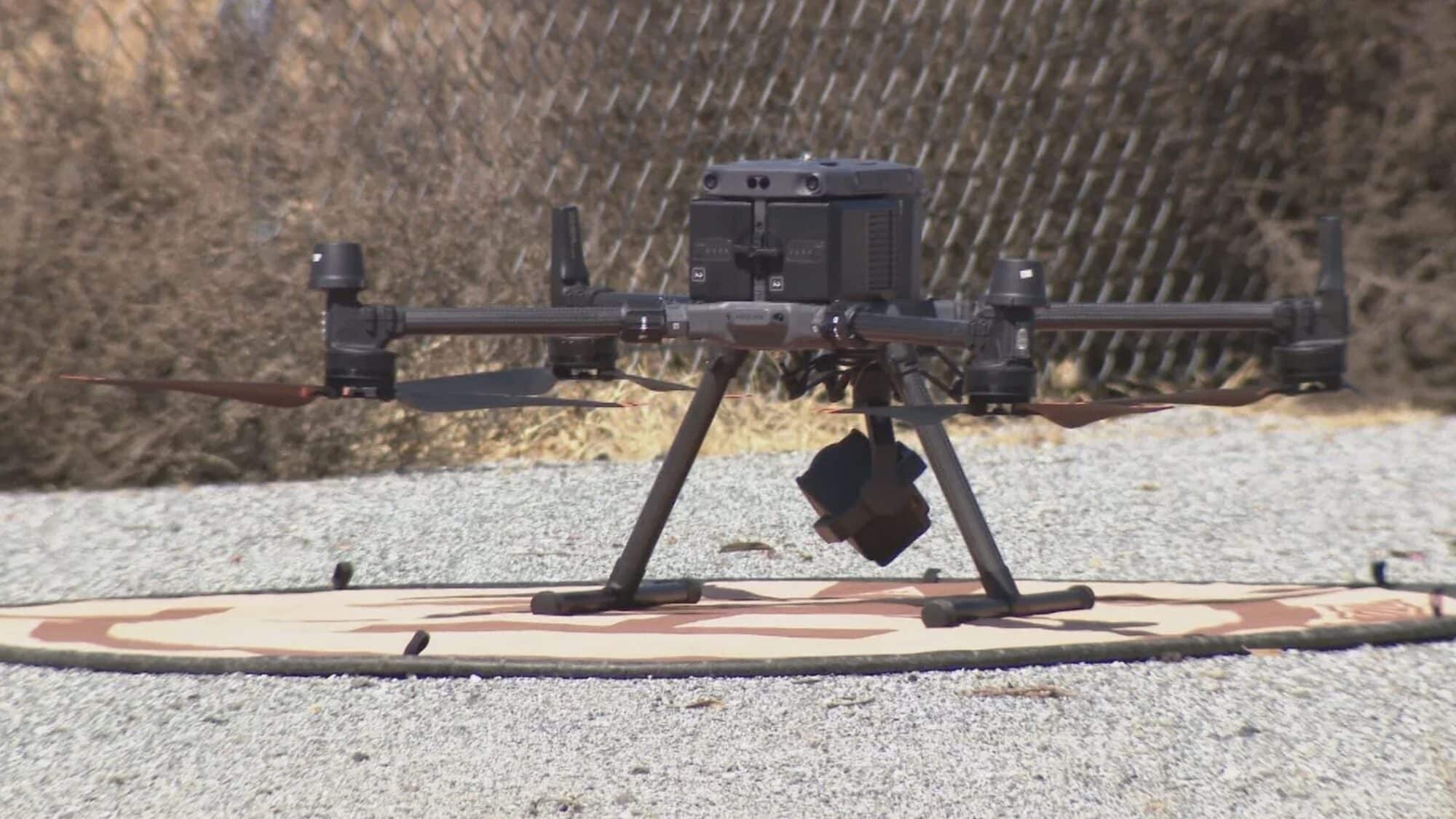 An inspection drone rests on a landing pad on gravel near a fence.