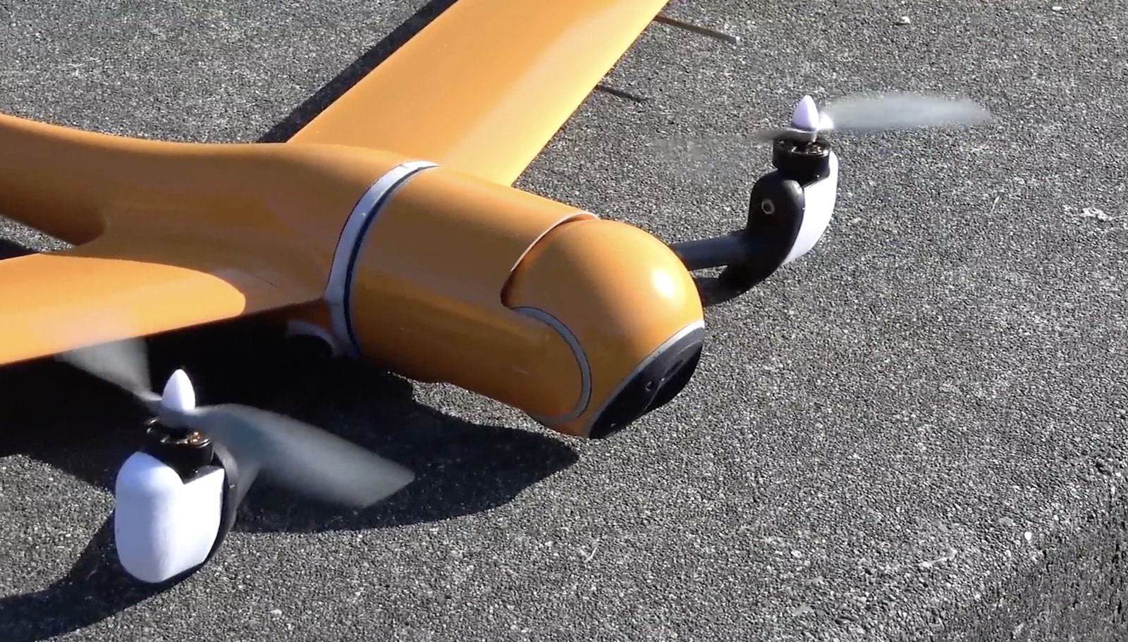 Tan fixed-wing tilt-rotor drone sitting on asphalt with propellers spinning.