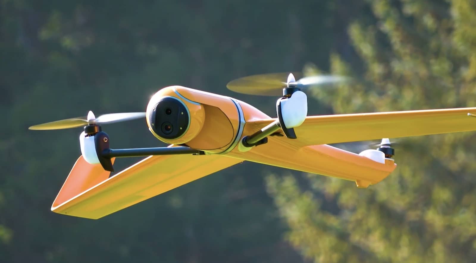A yellow tilt-rotor VTOL aircraft banks above a wooded hillside.