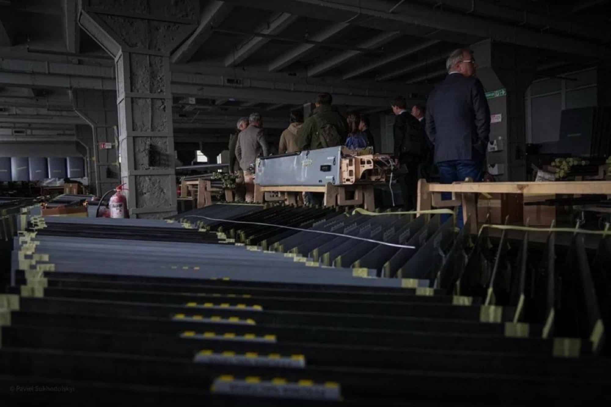 Rows of dark drone airframes crated on a warehouse floor as officials tour.