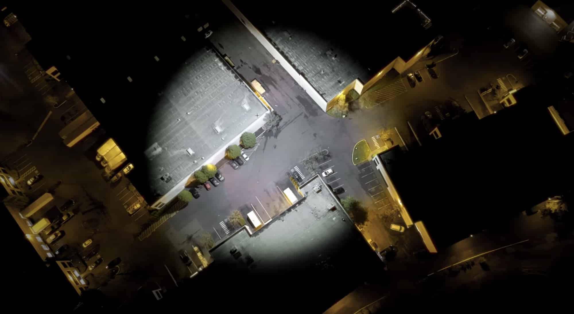 A drone-mounted floodlight throws a bright circle across a dark industrial yard.