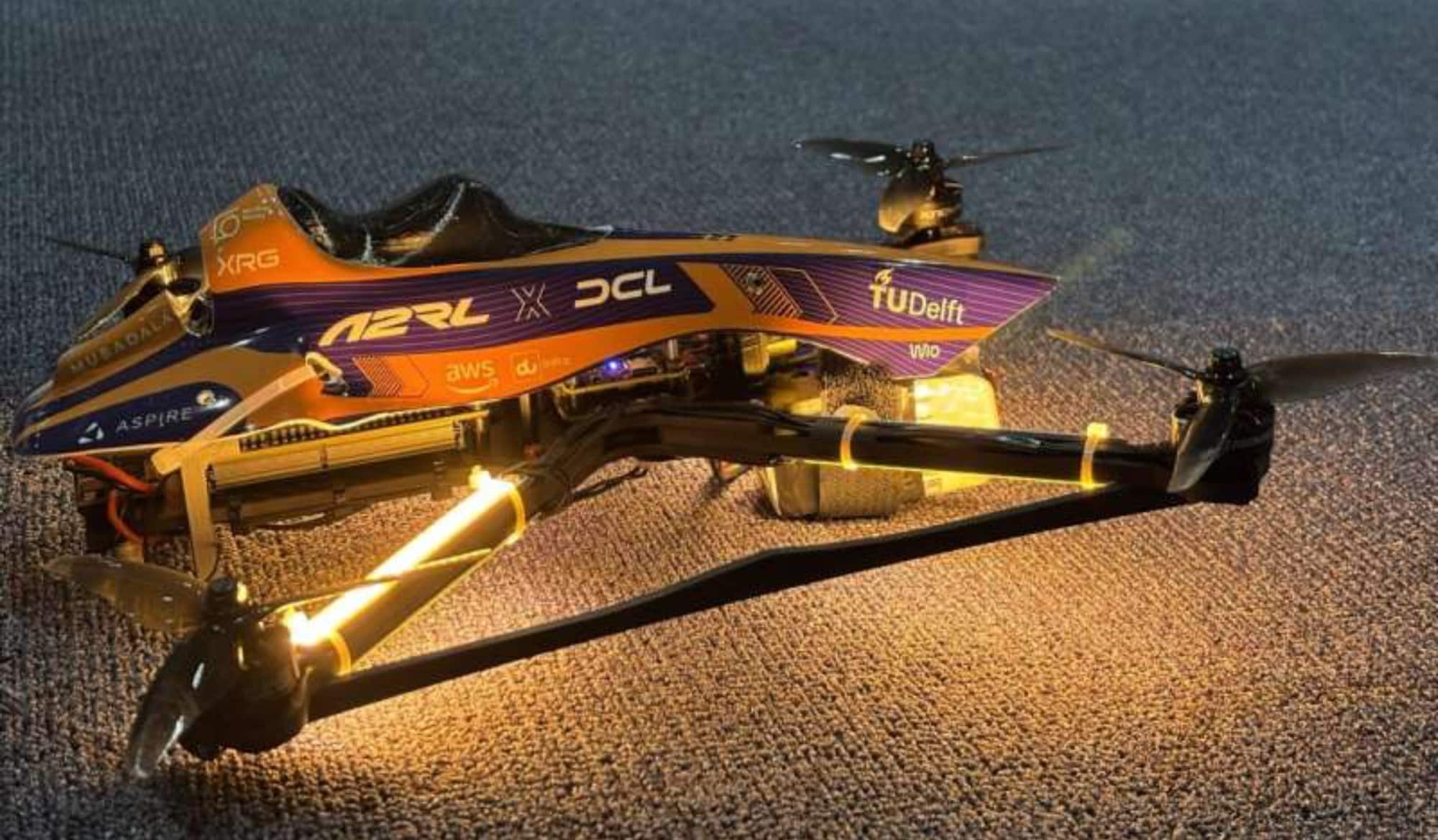 Orange and purple racing quadcopter glowing on gravel, wearing A2RL and TU Delft livery.