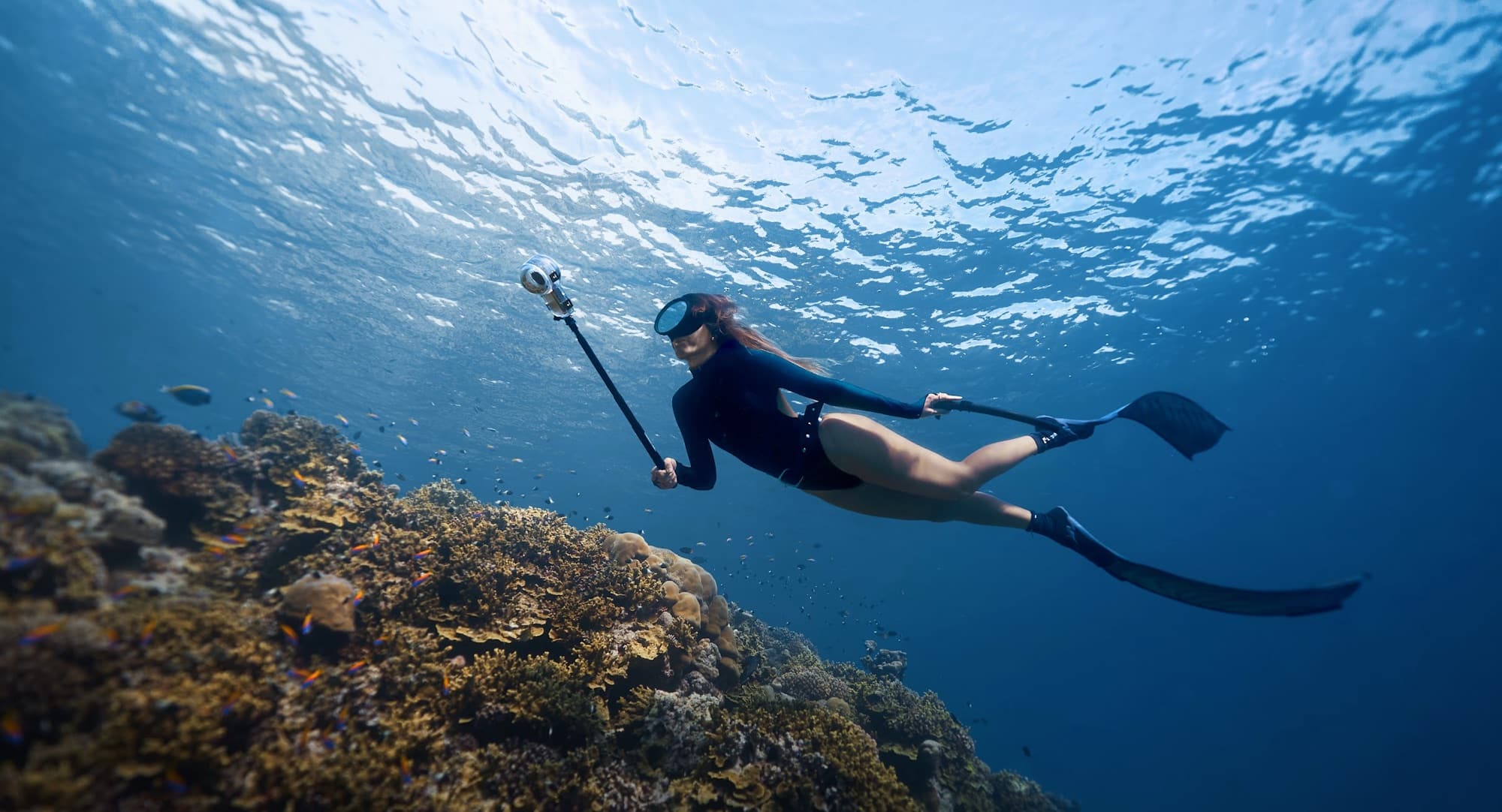 A freediver glides over a reef holding the camera on an invisible selfie stick.