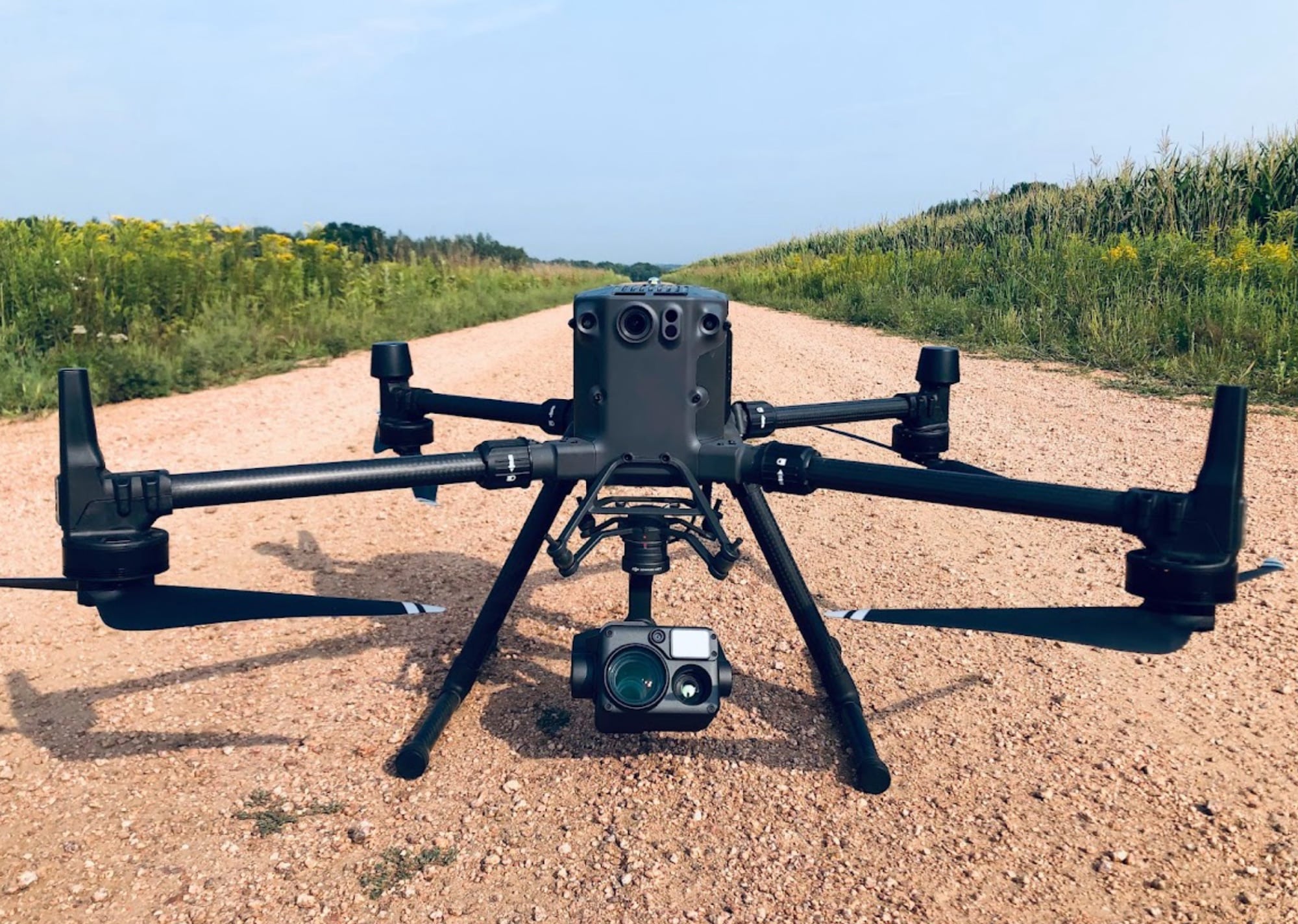 A Matrice quadcopter with a thermal gimbal sits on a gravel track by cornfields.