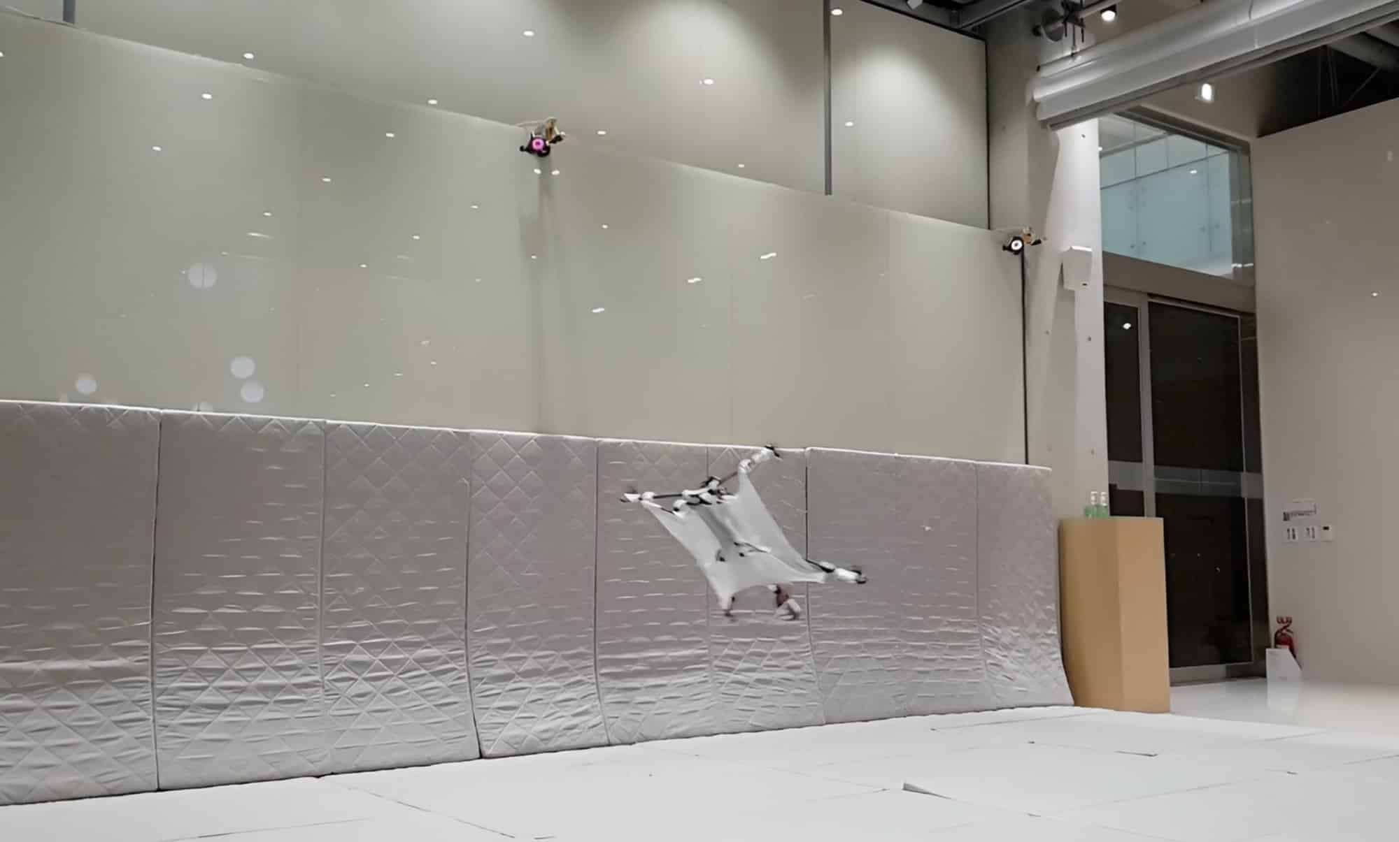 A winged test drone brakes in mid-air inside a padded indoor flight lab.