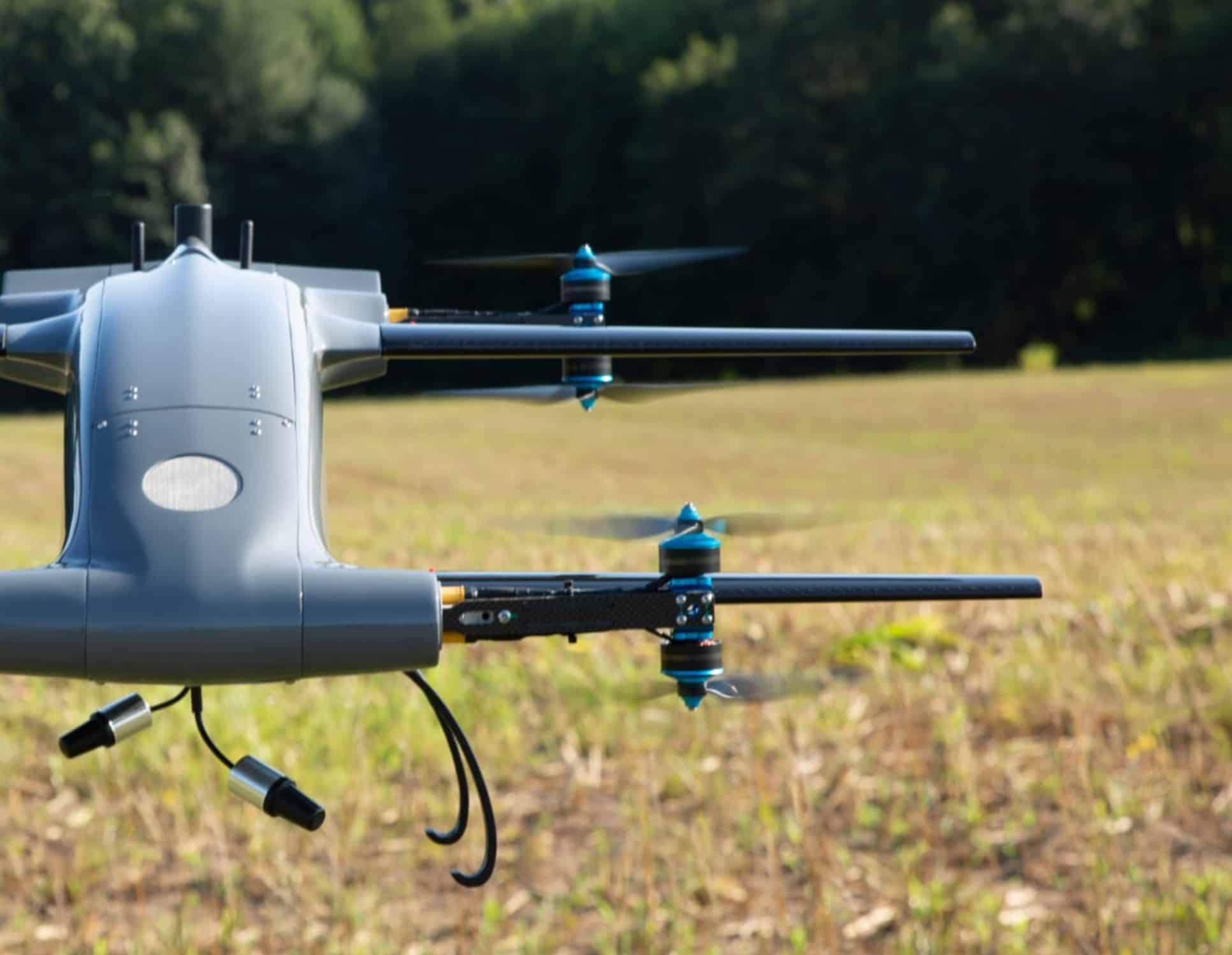 A pale blue Blueflite cargo VTOL drone flies low across a grass field.