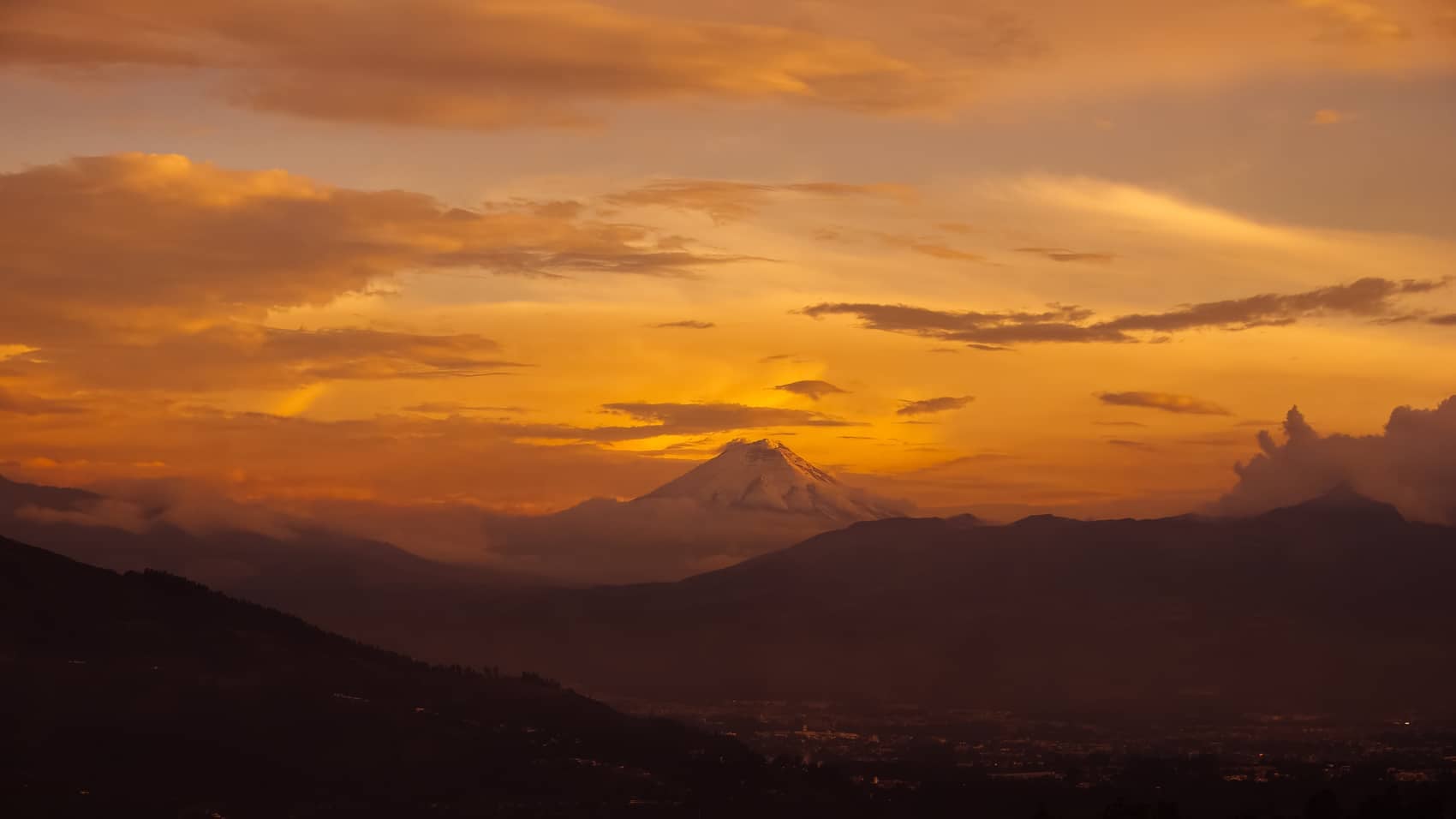 Aerial sunset over layered ridges with a snowcapped volcano against golden cloud.