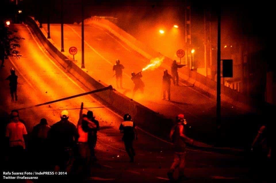 Protesters silhouetted on an orange-lit highway underpass during a night confrontation.