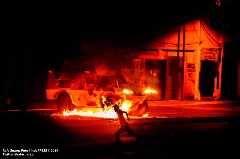 A vehicle burns in the dark as a lone figure walks past the flames.