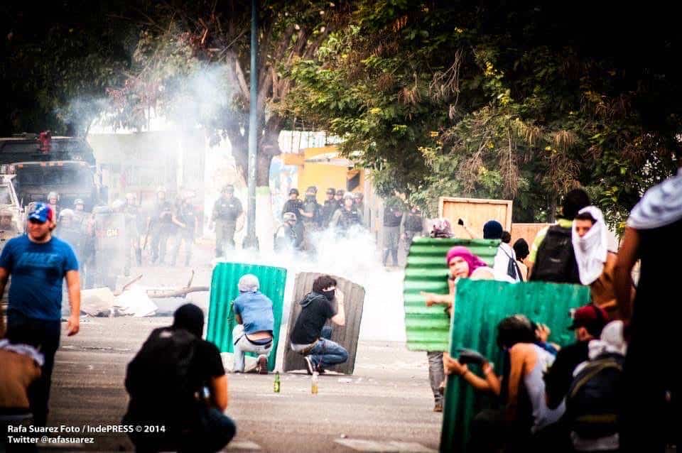 Demonstrators shelter behind green metal barricades as tear gas drifts across the road.