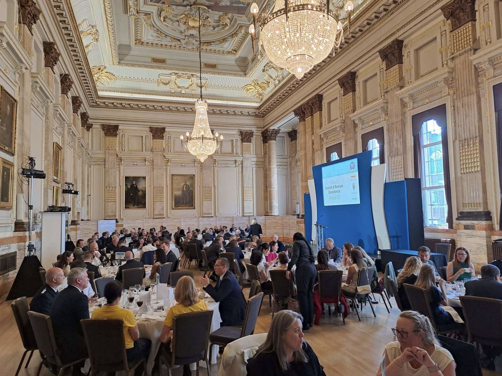Guests fill round tables beneath chandeliers in an ornate awards banquet hall.