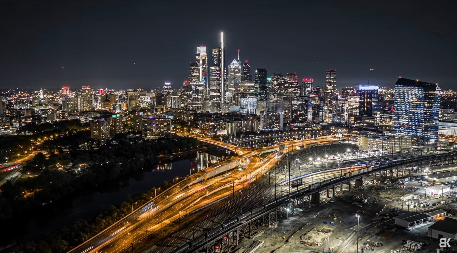 Night aerial of expressway light trails curving beside a river toward the skyline.